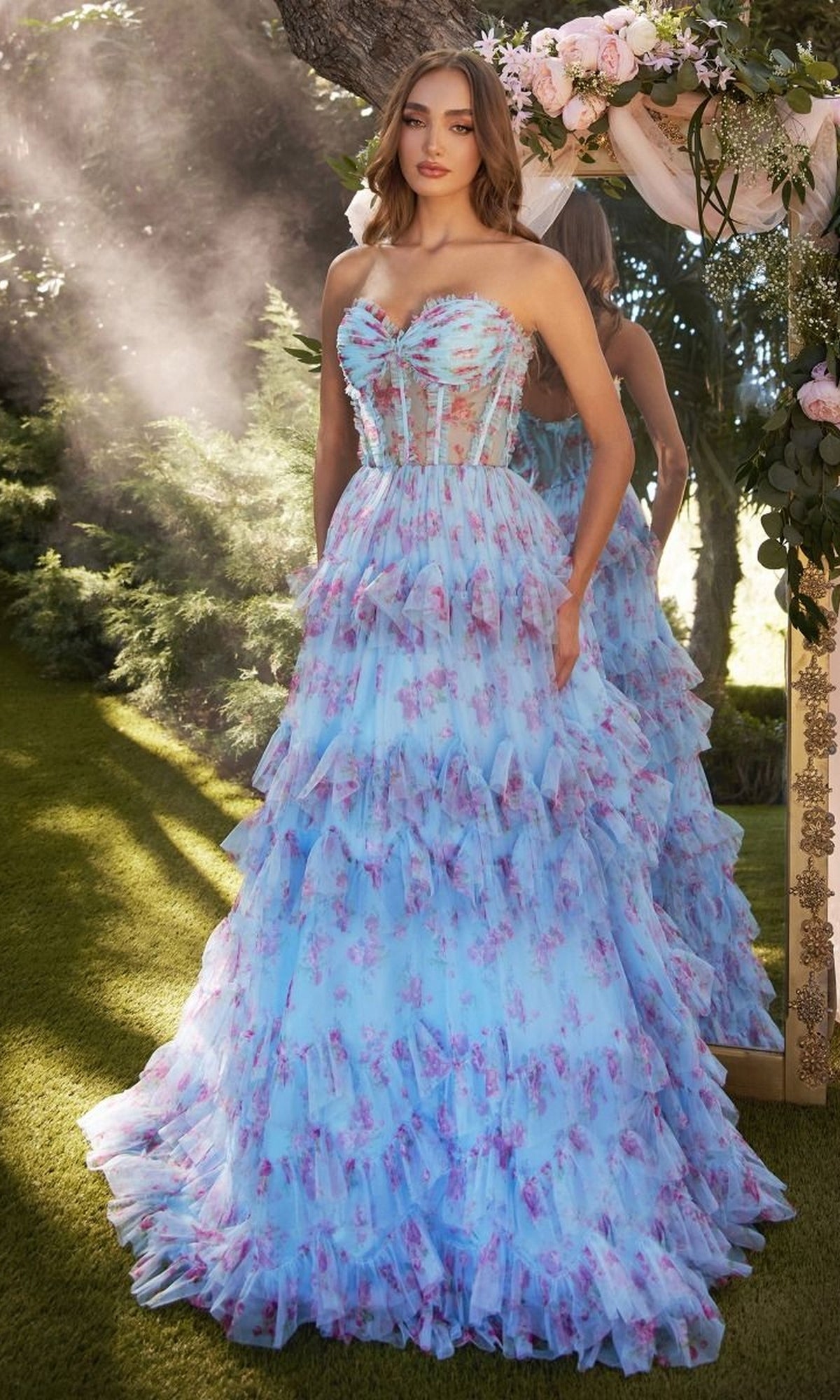 A woman in the Andrea and Leo A1334 strapless print prom dress with ruffles and a pink floral pattern stands outdoors by a mirror under a flower-adorned arch.
