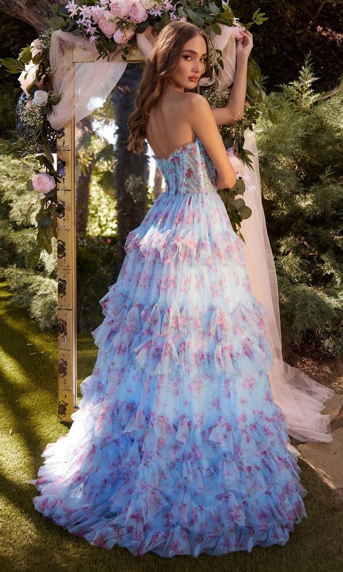 A woman in the Andrea and Leo A1334 Strapless Print Prom Dress with ruffled blue layers and pink floral patterns stands outdoors, gazing over her shoulder beside a flower- and greenery-adorned mirror.