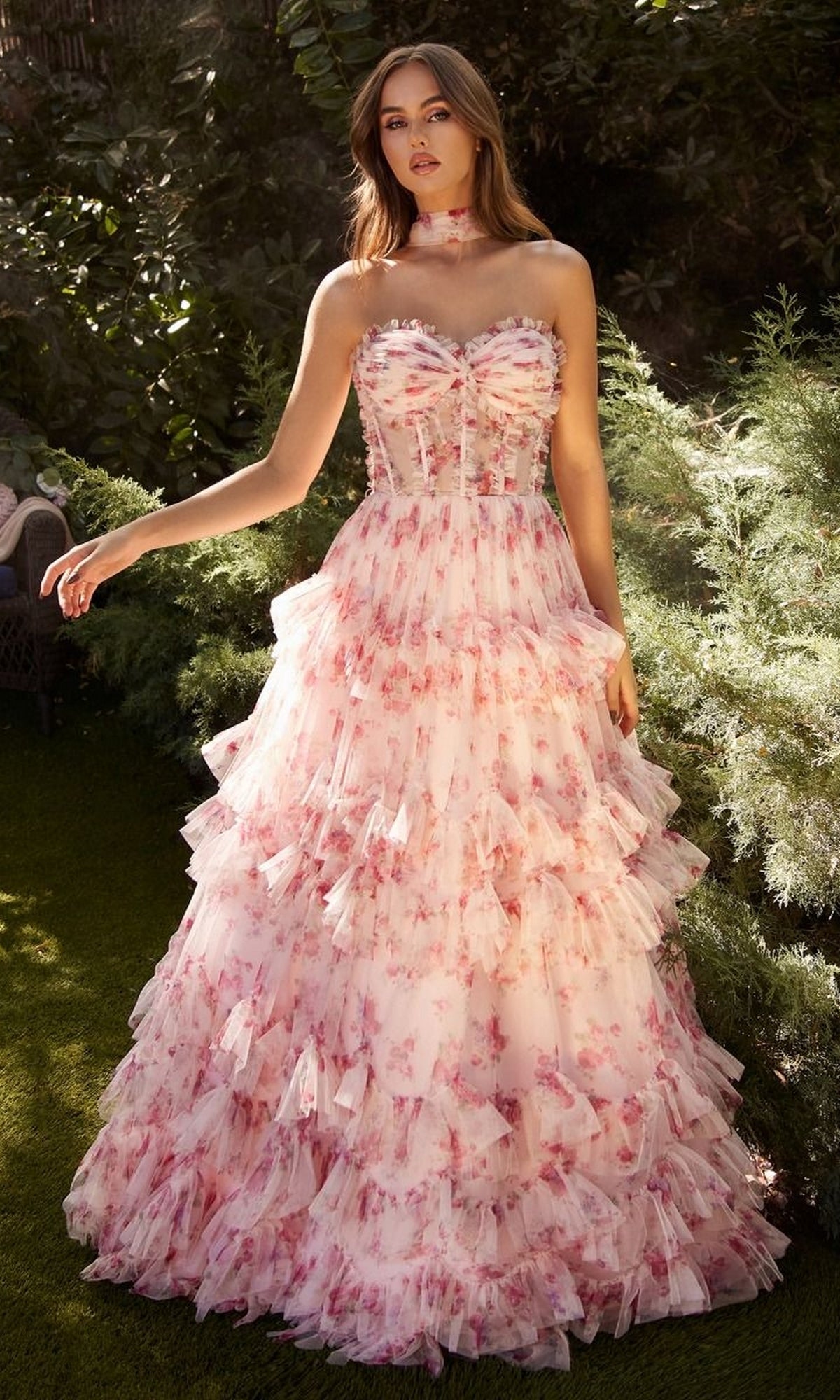A woman stands outdoors surrounded by greenery, wearing the Andrea and Leo A1334 Strapless Print Prom Dress in pink and white with ruffles and a matching choker.