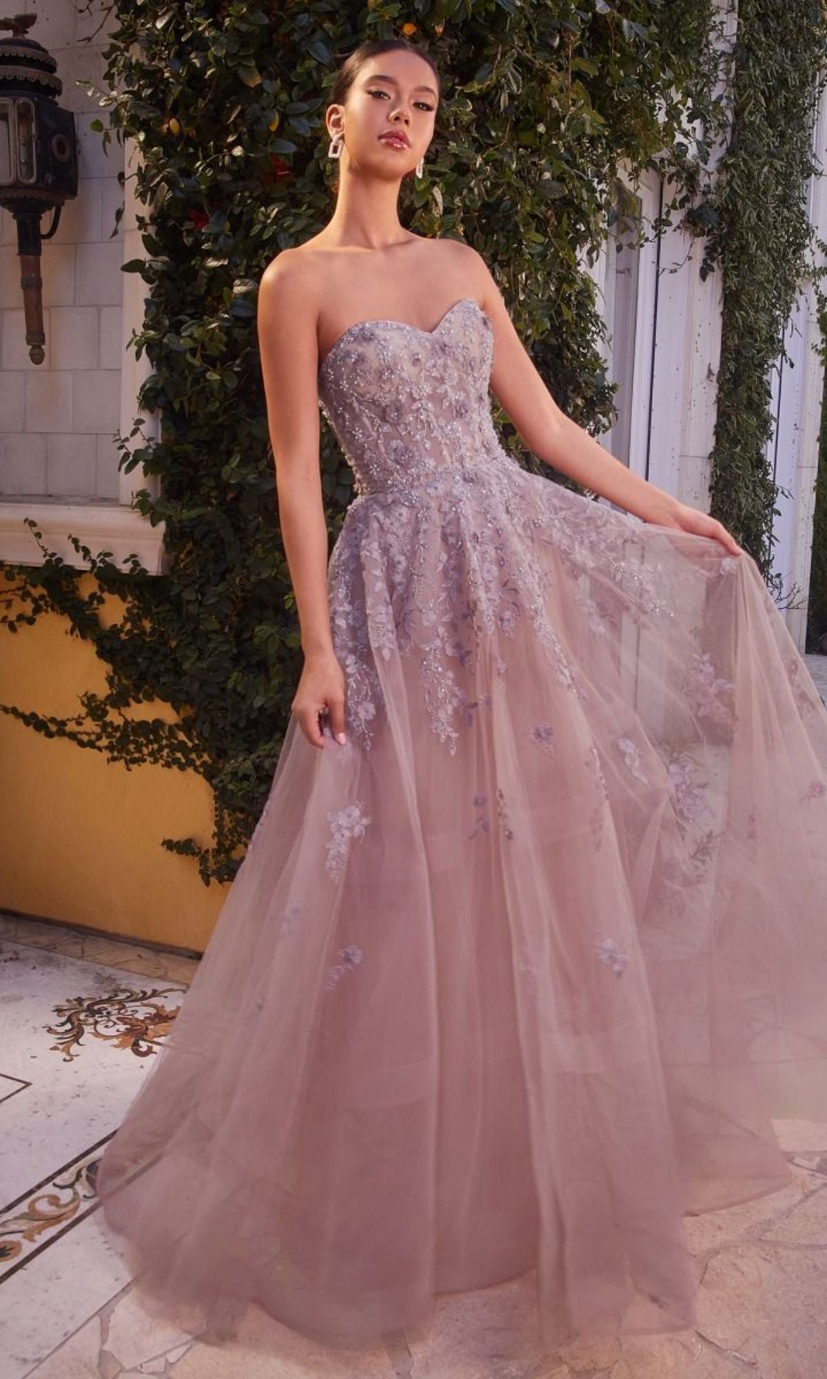 A woman wearing the Long Strapless Sweetheart A-Line Prom Dress A1348 in lavender with floral embroidery stands outdoors by a yellow wall and greenery.