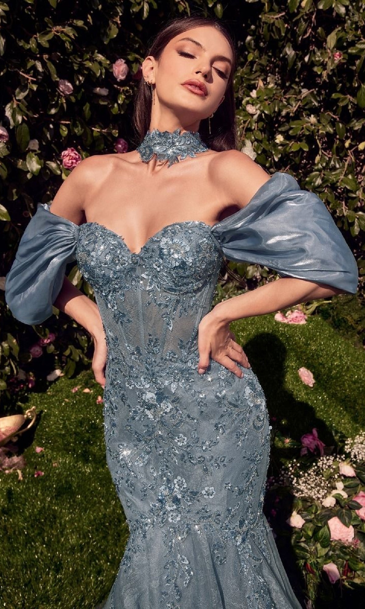 A woman poses outdoors in the Andrea & Leo A1354, a fitted light blue off-the-shoulder mermaid prom dress with floral embellishments, surrounded by lush greenery and blooming flowers.