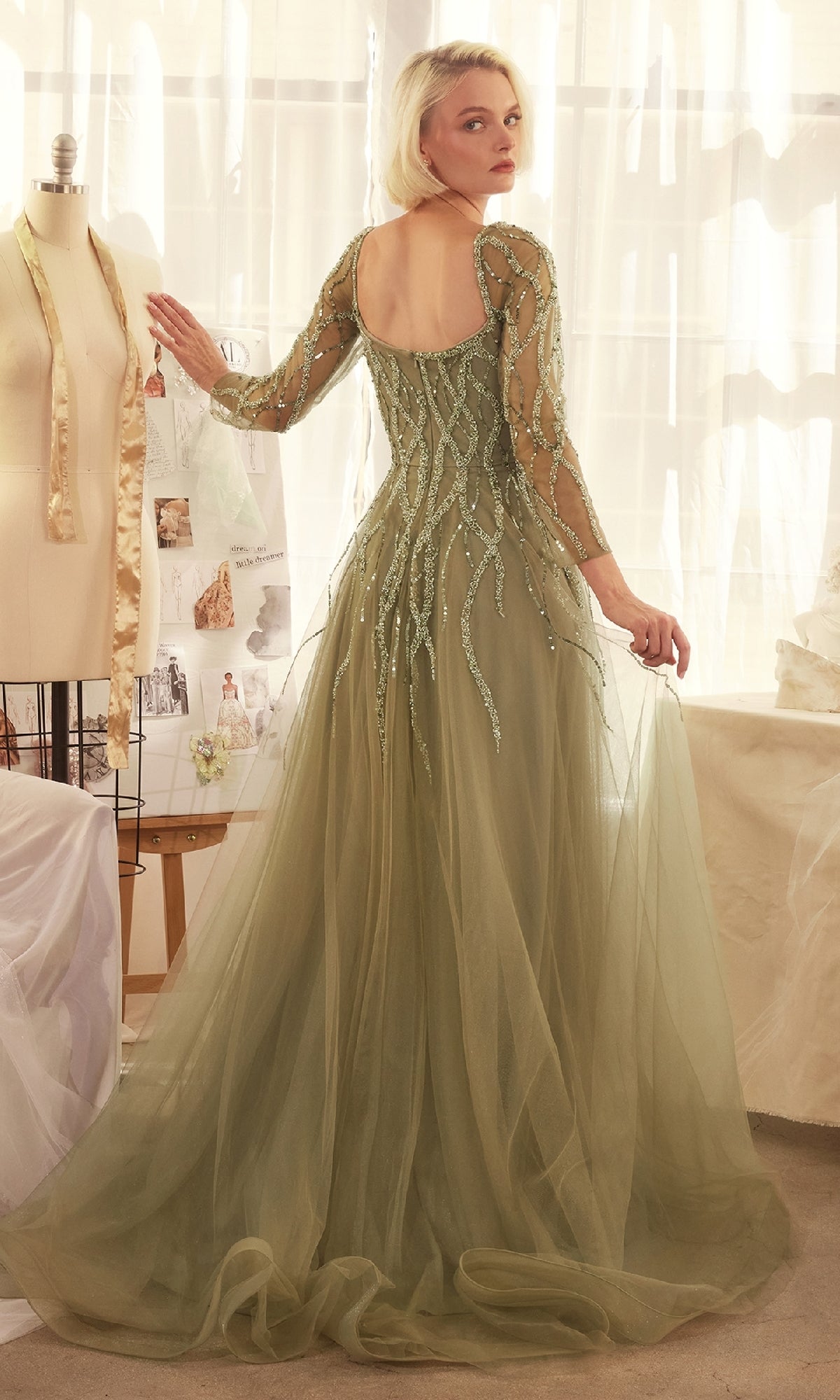 A woman wearing the Andrea & Leo A1367 Long Sleeve Prom Ball Gown in pale green with sheer beaded details stands in a sunlit room, looking over her shoulder.