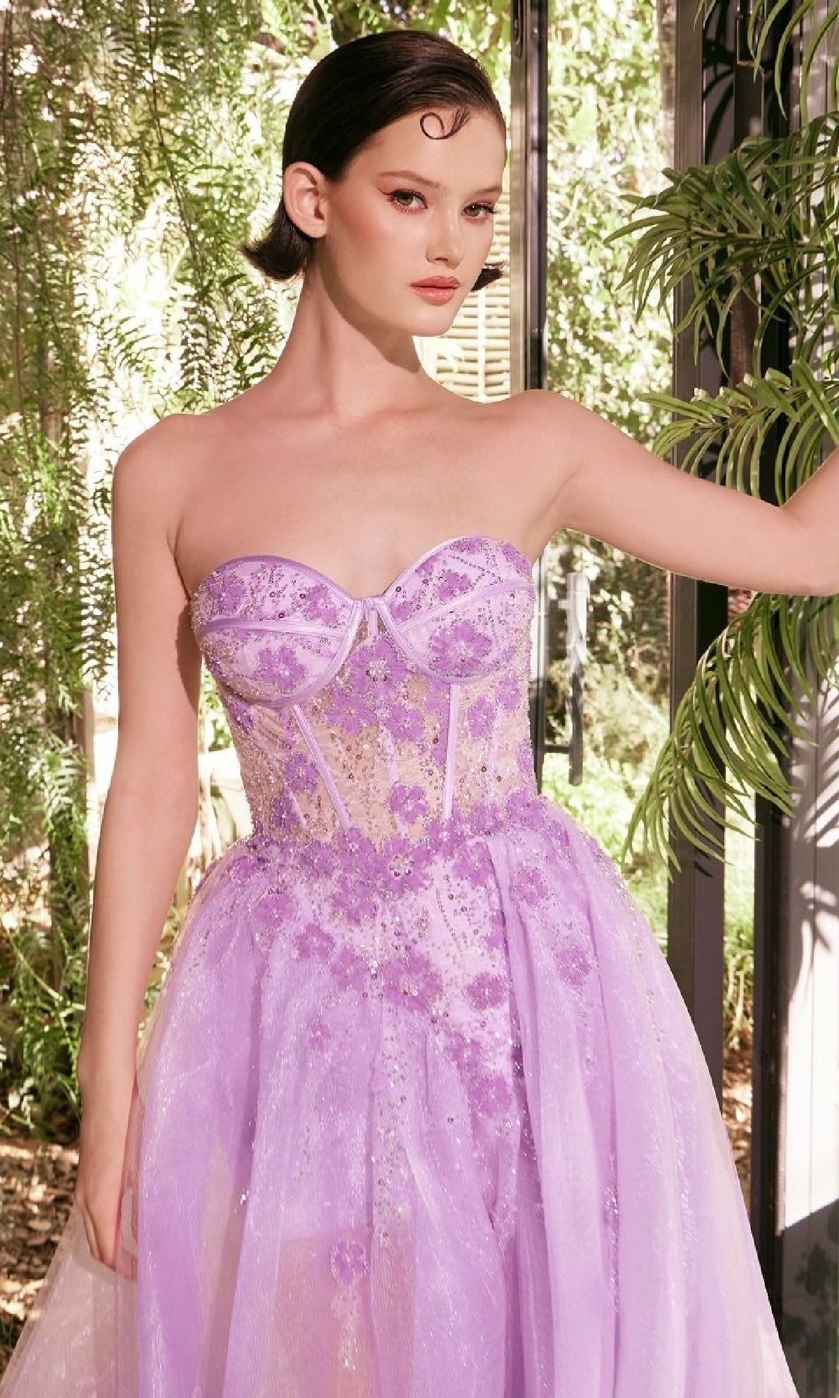 A woman stands indoors wearing the Andrea & Leo A1381 Strapless Corset Prom Gown, featuring a sweetheart neckline, beaded floral details, and a sheer lavender overlay. Sunlight and green plants appear in the background.