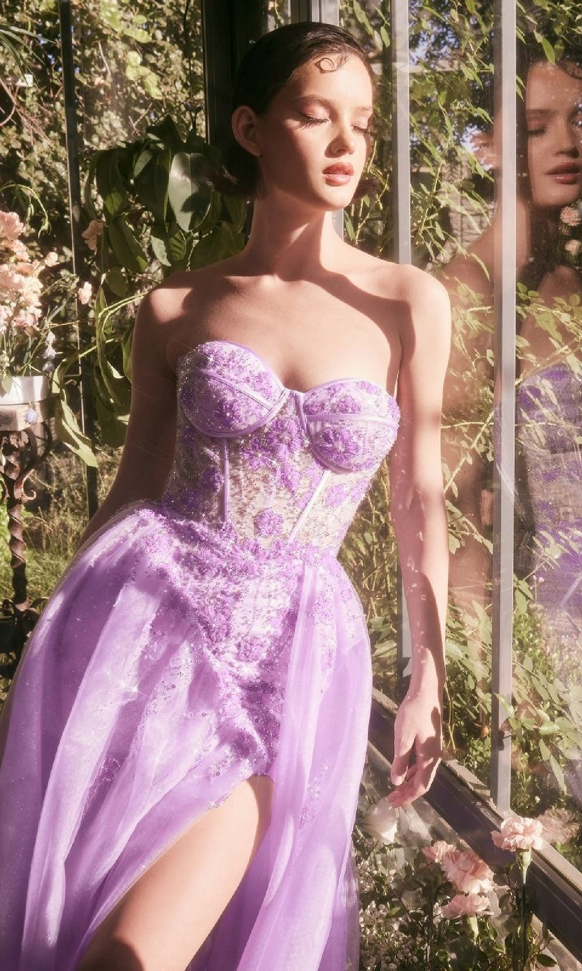 A woman in the Strapless Corset Prom Gown: Andrea & Leo A1381 stands by a sunlit window amid greenery and flowers. Beaded floral details add elegance to her lavender glittery dress as her reflection shimmers in the glass.