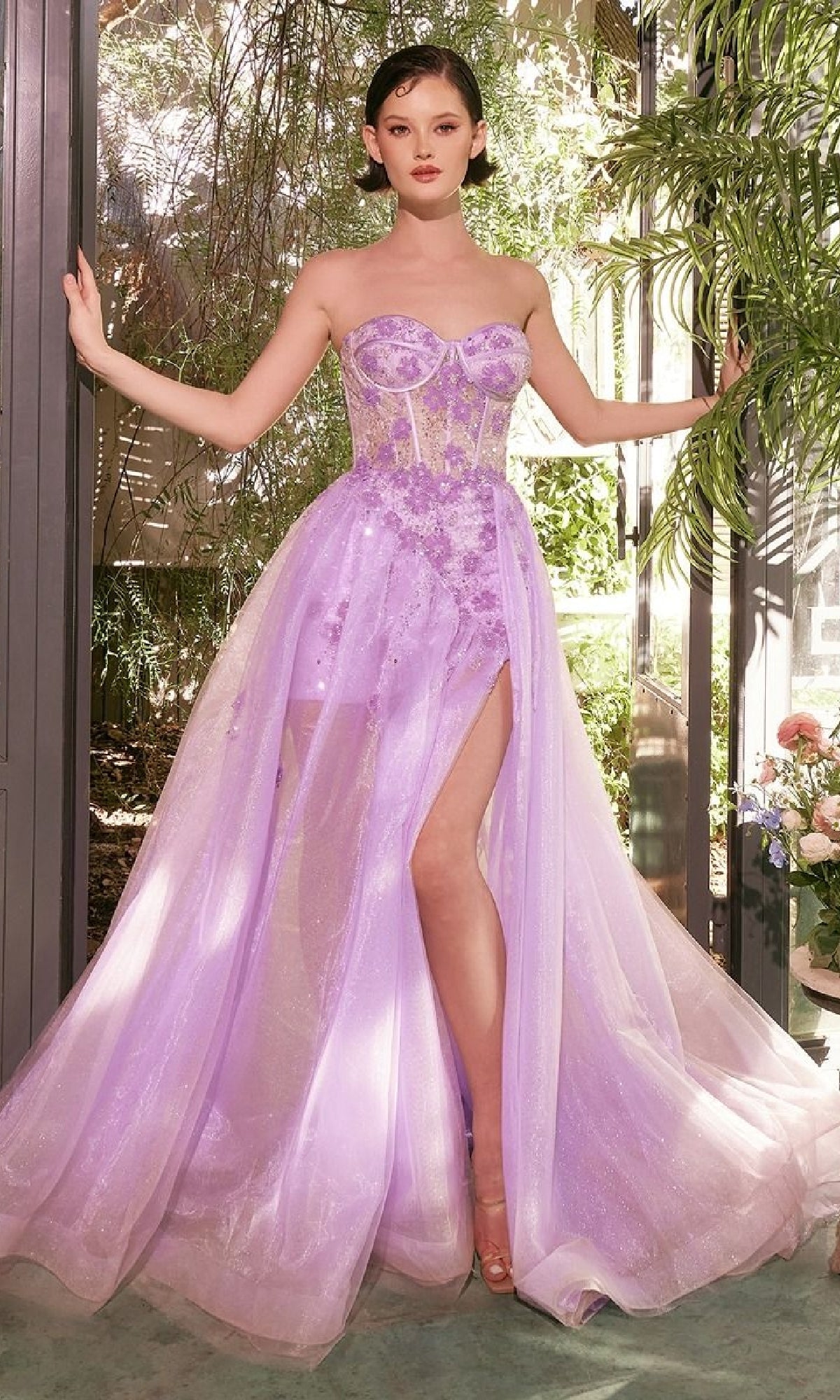A woman wearing the Strapless Corset Prom Gown: Andrea & Leo A1381 in lilac, featuring a strapless sweetheart neckline, sheer bodice with beaded floral accents, high slit, and tulle skirt stands indoors by windows and lush plants.
