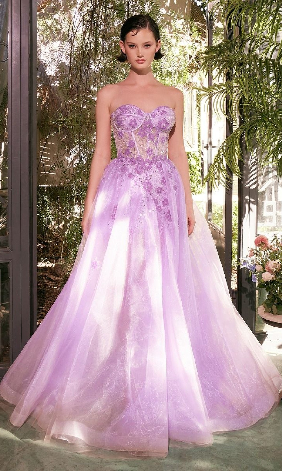 A woman stands indoors wearing the Andrea & Leo A1381 Strapless Corset Prom Gown in lavender, featuring sheer panels and beaded floral embellishments, surrounded by lush green plants and flowers.