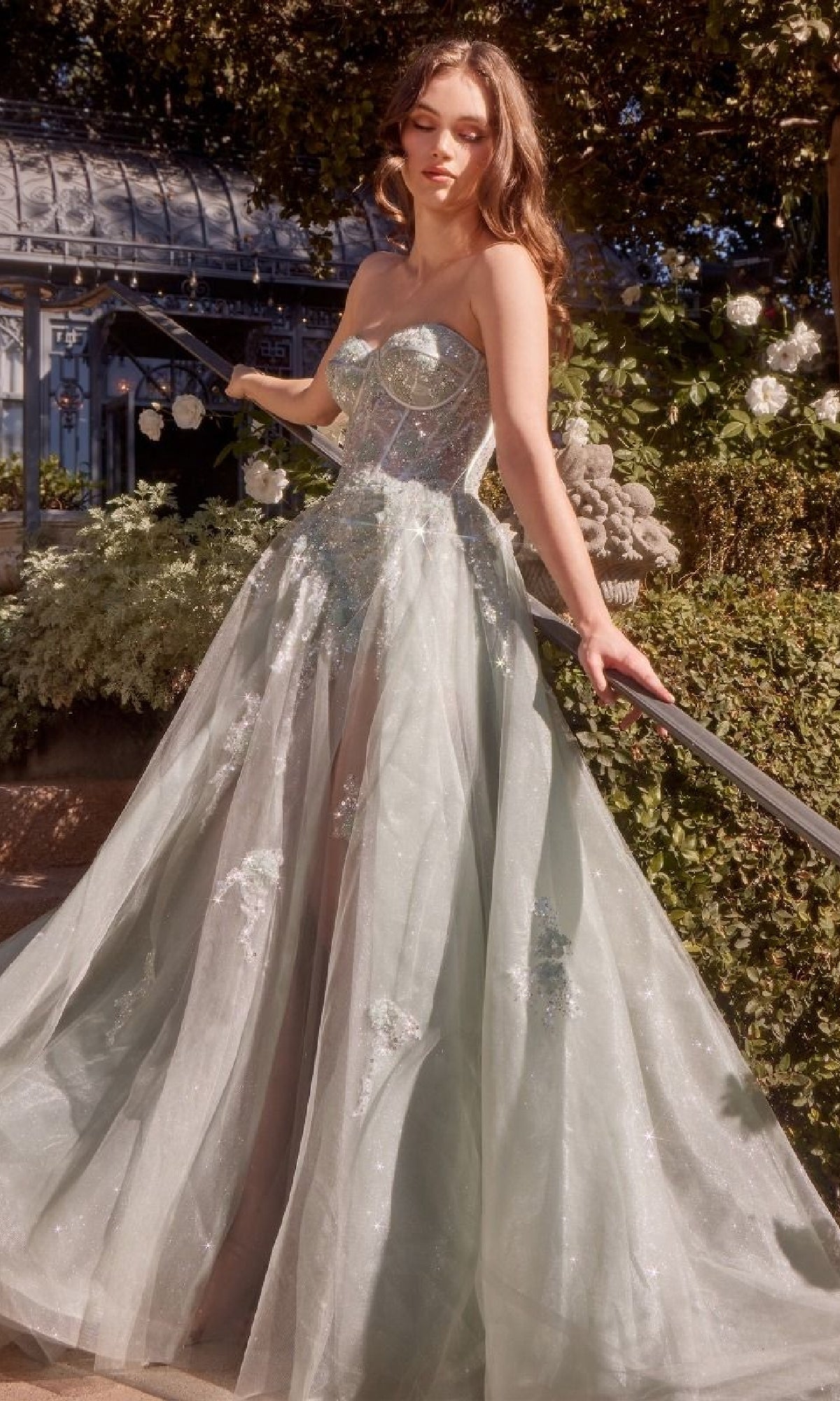 A woman wearing the Strapless Corset Prom Gown Andrea & Leo A1381, featuring a sweetheart neckline, beaded floral details, and a full tulle skirt, stands outdoors near flowers and greenery.