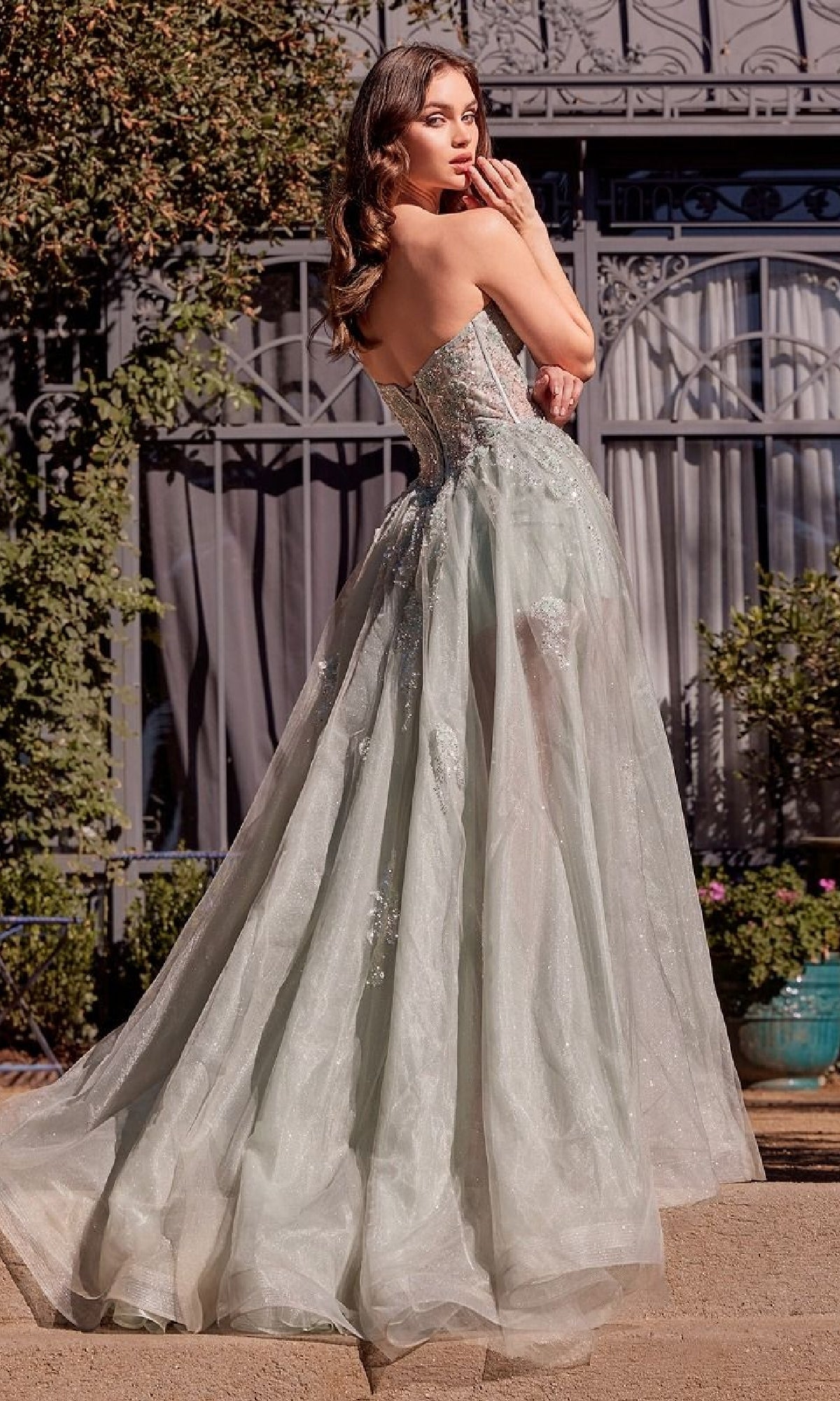 A woman in the Strapless Corset Prom Gown: Andrea & Leo A1381, featuring a sweetheart neckline, beaded floral embellishments, and a sheer flowing skirt, stands outdoors and glances over her shoulder.