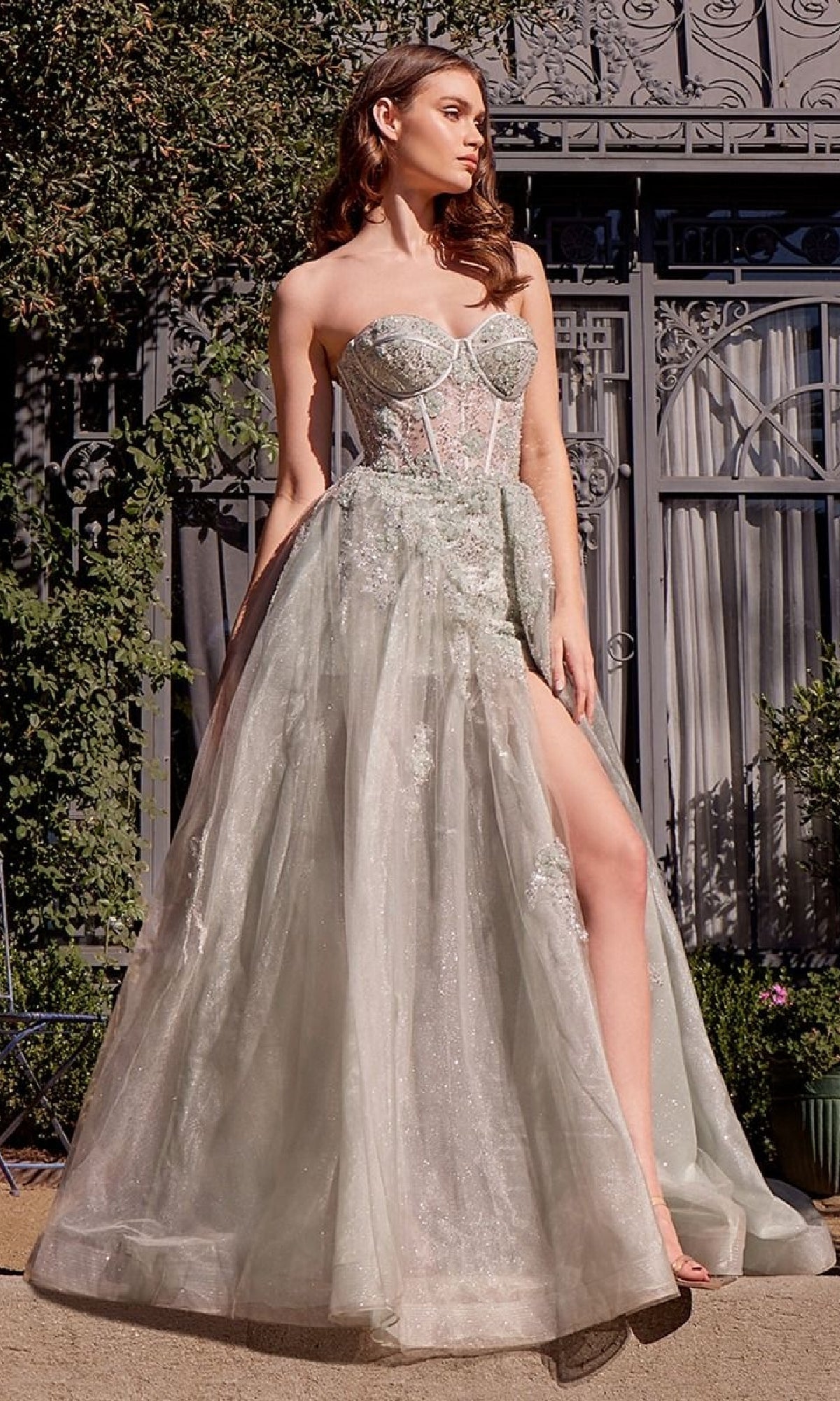 A woman poses outdoors in the Andrea & Leo A1381 Strapless Corset Prom Gown, featuring a sweetheart neckline, beaded floral accents, sheer bodice, high slit, and sparkling details before an ornate gate and lush greenery.
