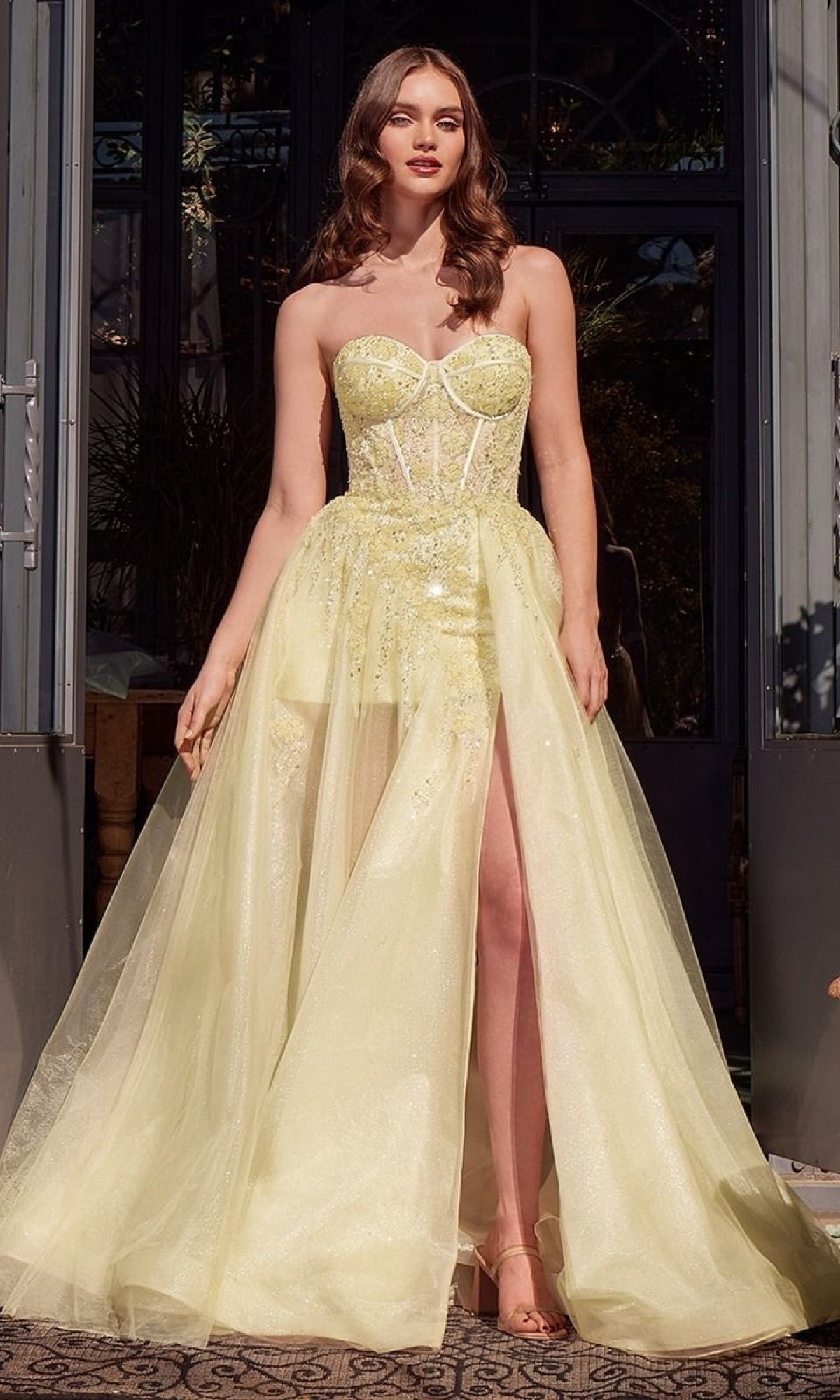 A woman poses outdoors in front of glass doors wearing the Strapless Corset Prom Gown: Andrea & Leo A1381, a light yellow sweetheart dress with sheer overlay, high slit, and beaded floral embellishments.