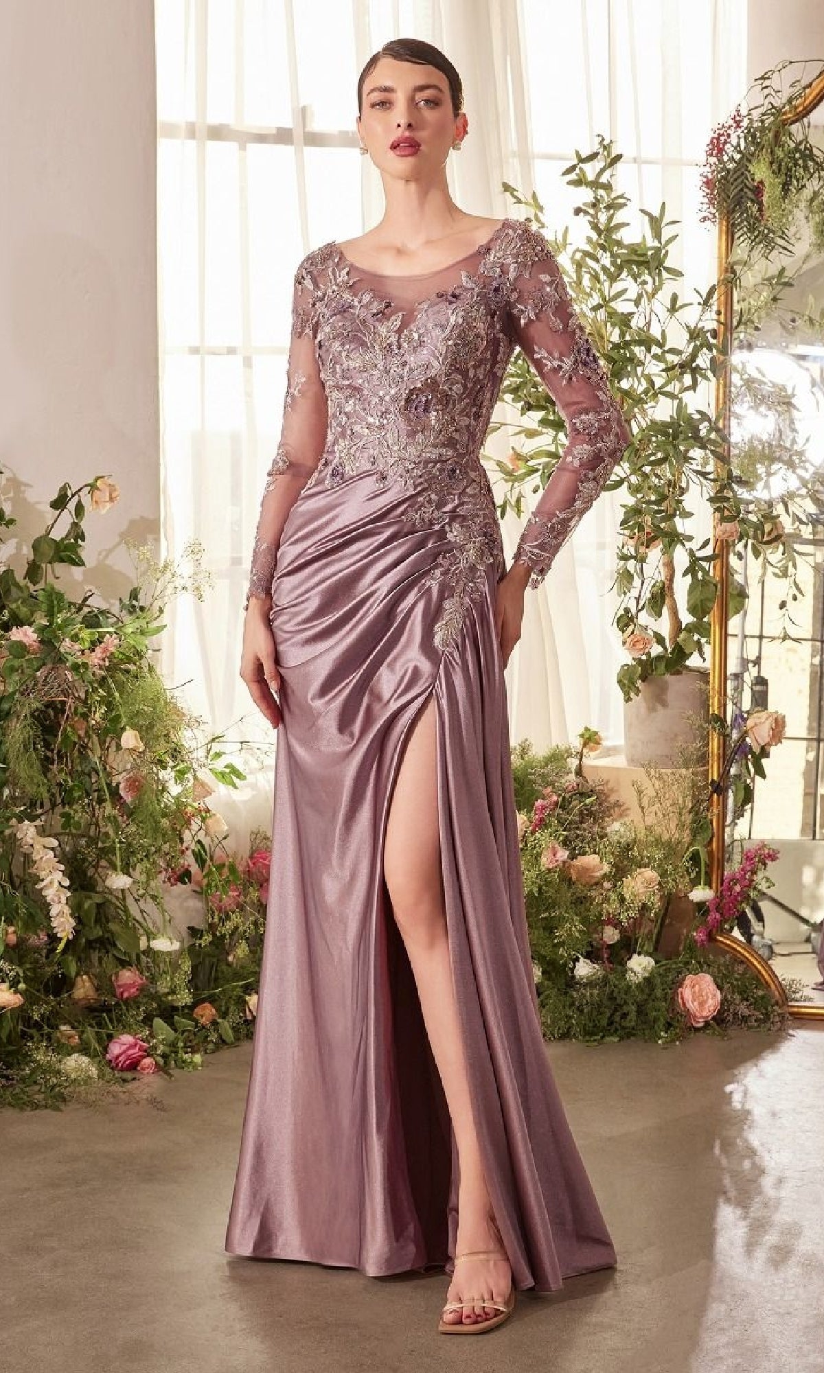 A woman wearing the Andrea and Leo A1403 long formal dress, featuring mauve satin with ruched skirt, floral lace bodice, sheer embroidered sleeves, and a thigh-high slit, stands indoors among floral arrangements.
