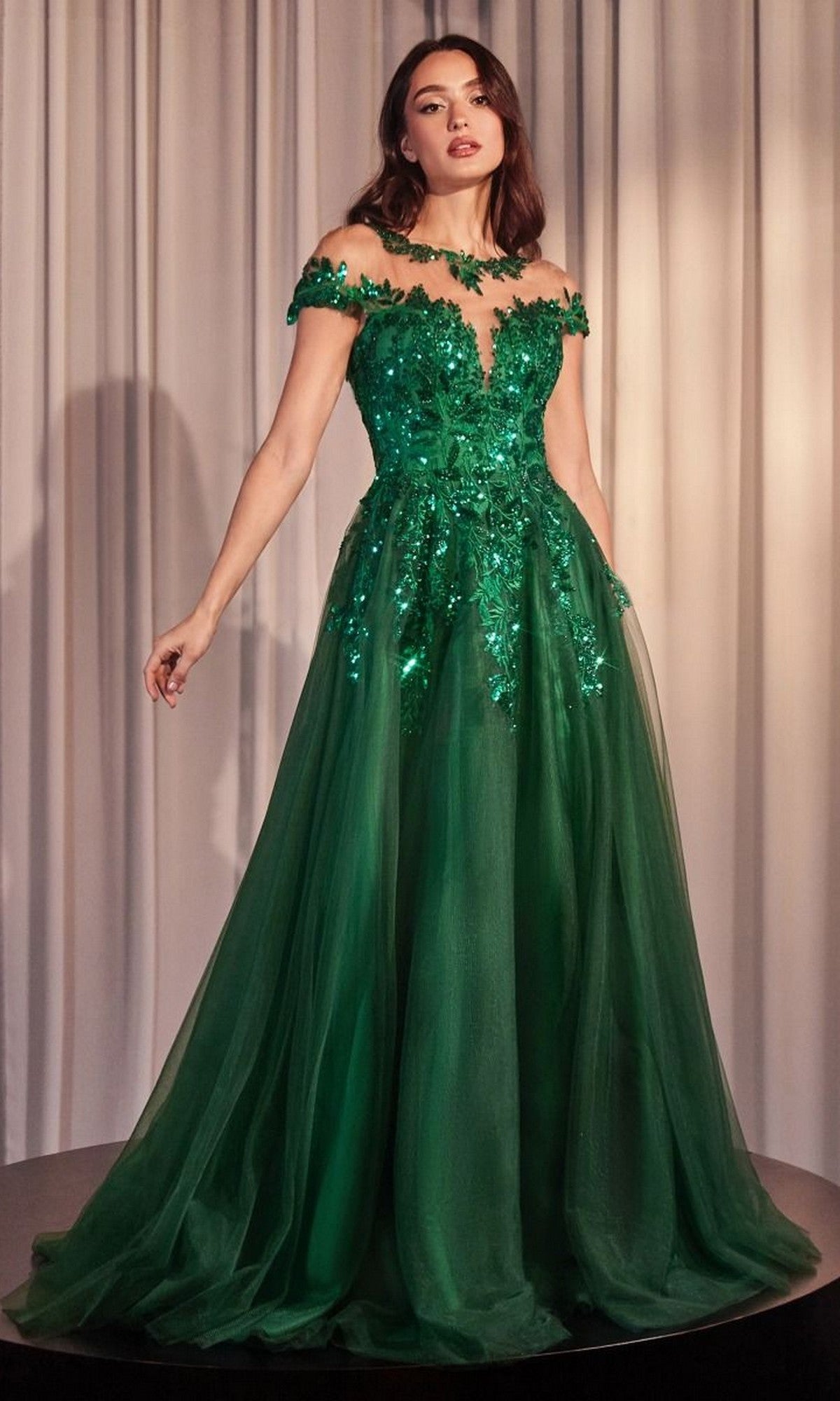 A woman stands in front of light-colored curtains wearing the Andrea and Leo A1475 emerald long prom dress, featuring an off-the-shoulder design with sequins and floral appliques.