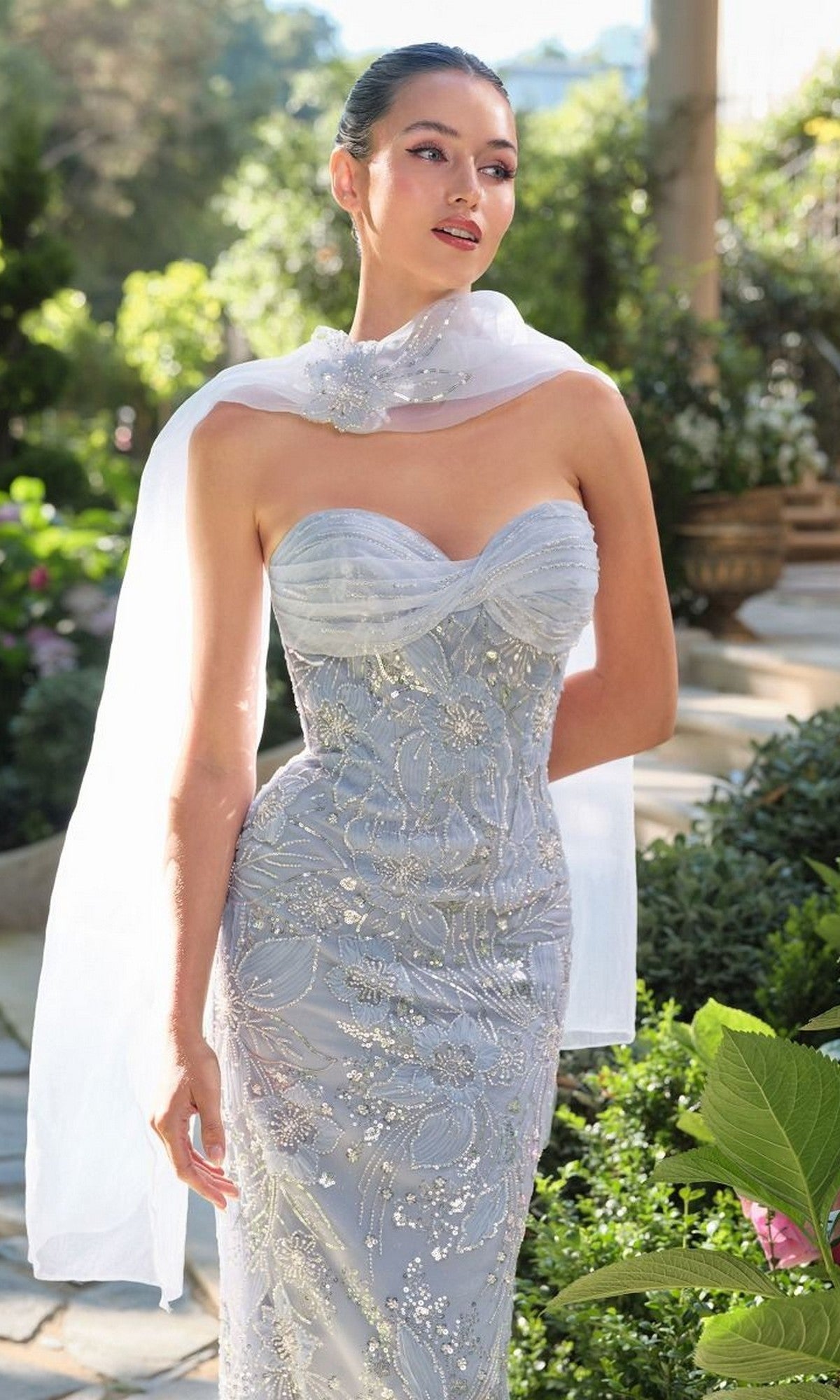 A woman in the Andrea and Leo A1484 Strapless Beaded Prom Gown with a sheer white scarf stands outdoors in a lush garden.