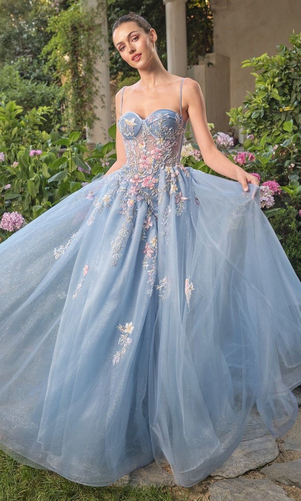 A woman stands outdoors in a garden wearing the Long Blue Prom Ball Gown: Andrea and Leo A1490, a blue floor-length dress with floral embroidery and thin straps, holding the skirt out to each side.