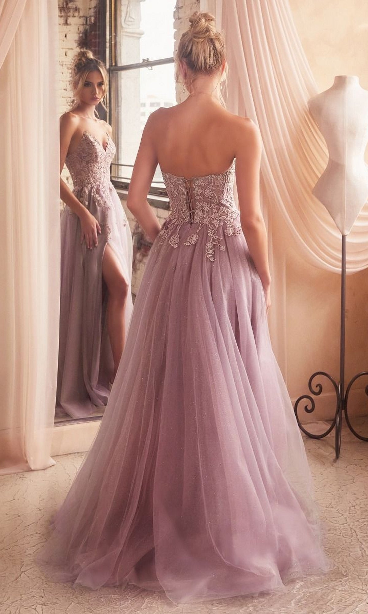 A woman in a Strapless Long Glitter-Tulle Prom Ball Gown C148 with an embroidered-lace bodice and tulle skirt stands before a mirror in a softly lit room with draped curtains and a dress form.