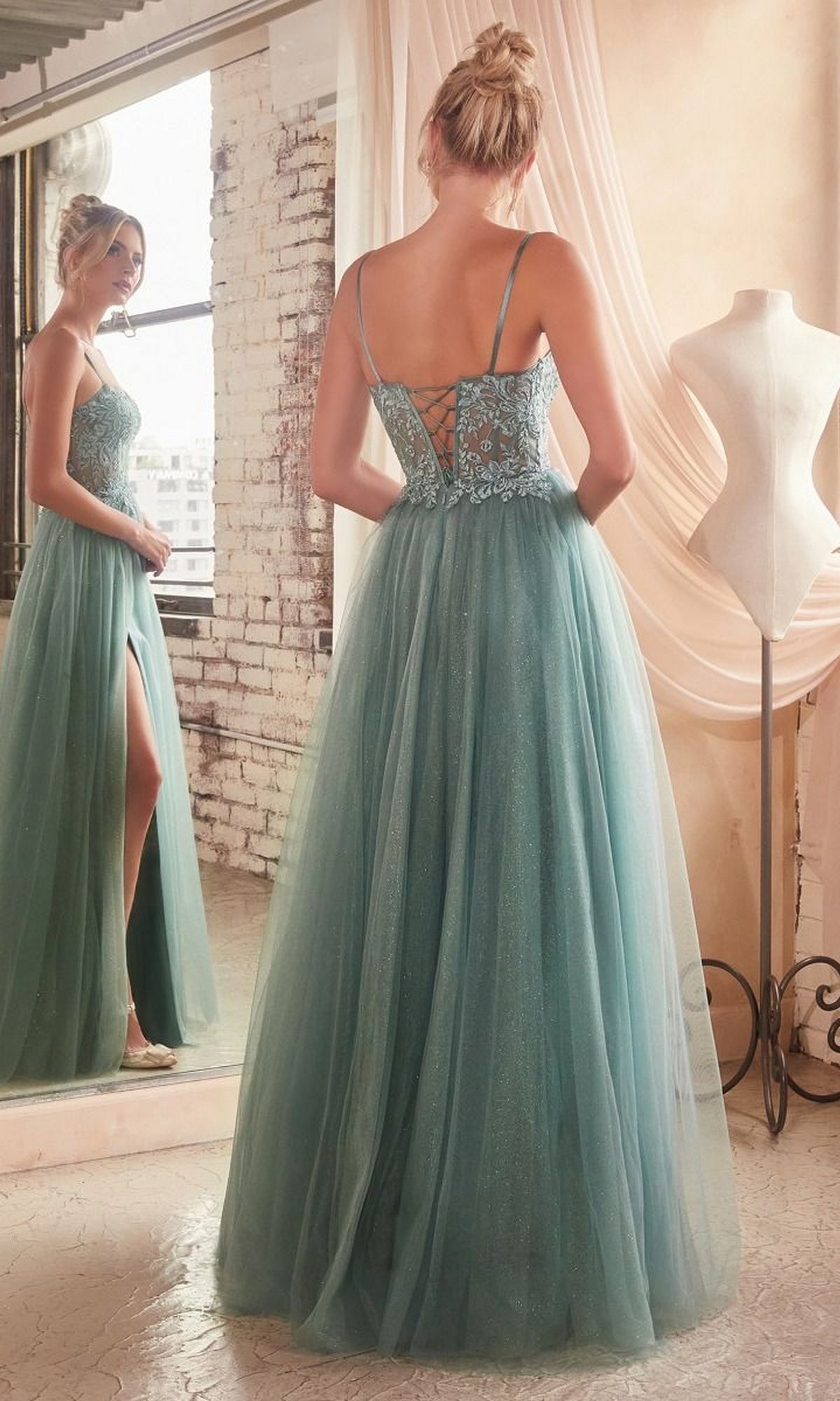 A woman in the Sheer-Waist Long Glitter-Tulle Prom Dress C150, featuring a lace-up back and thigh-high slit, stands by a window and dress form, gazing at her reflection in the mirror.