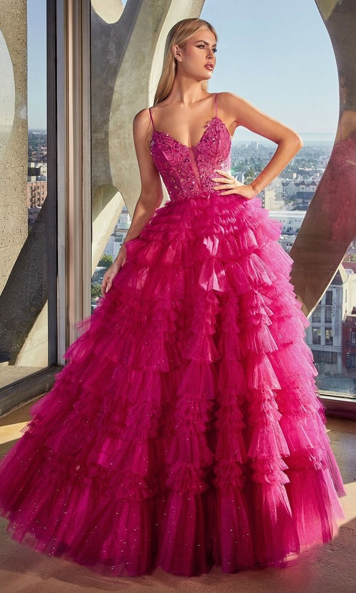 A woman stands indoors in the Sheer-Corset Long Glitter-Tulle Prom Ball Gown C152—bright pink, ruffled, with a fitted bodice and thin straps—gazing out a window at the cityscape beyond.
