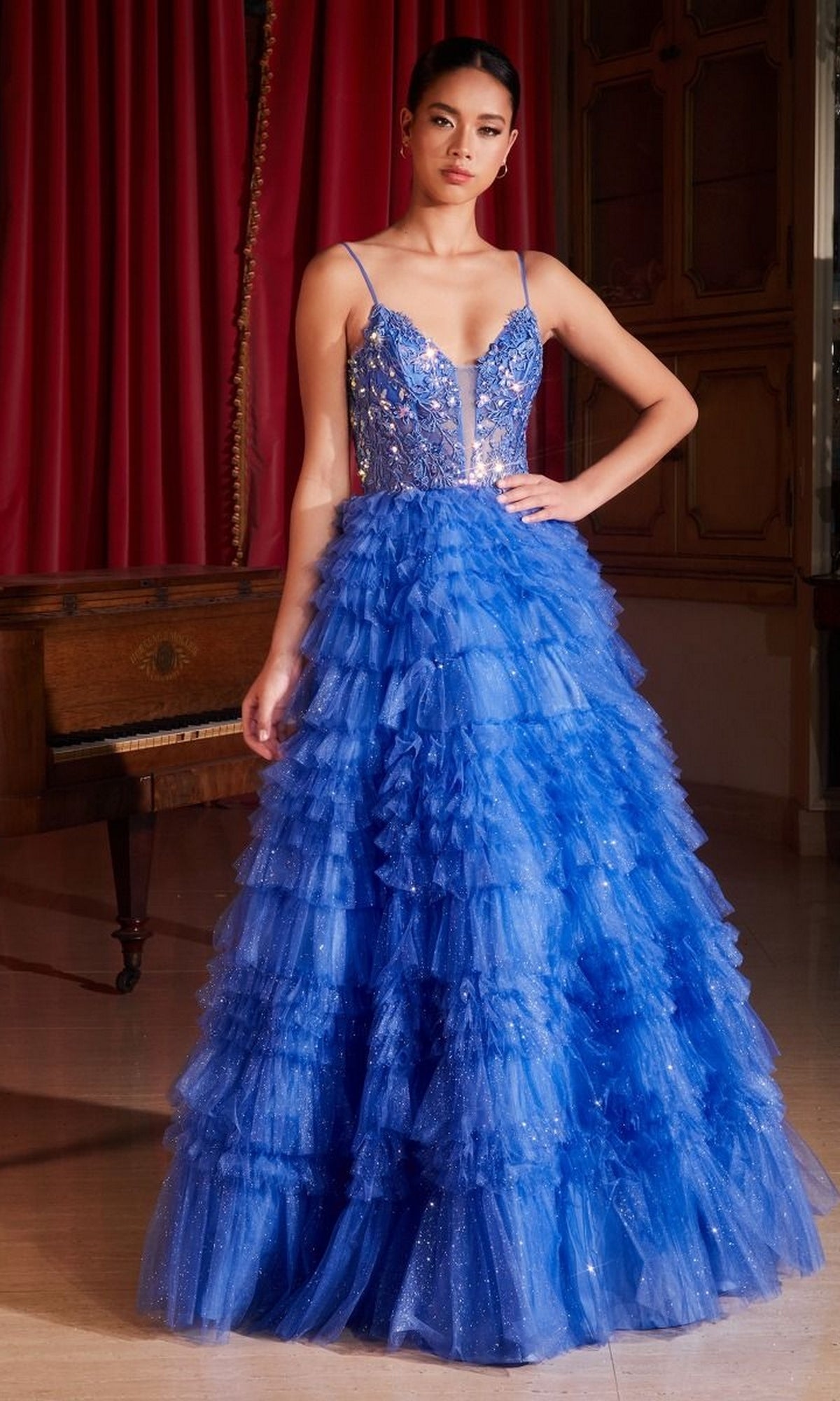 A woman stands indoors wearing the Sheer-Corset Long Glitter-Tulle Prom Ball Gown C152, featuring a fitted embellished bodice, thin straps, and elegant ruffled layers that flow gracefully to the floor.