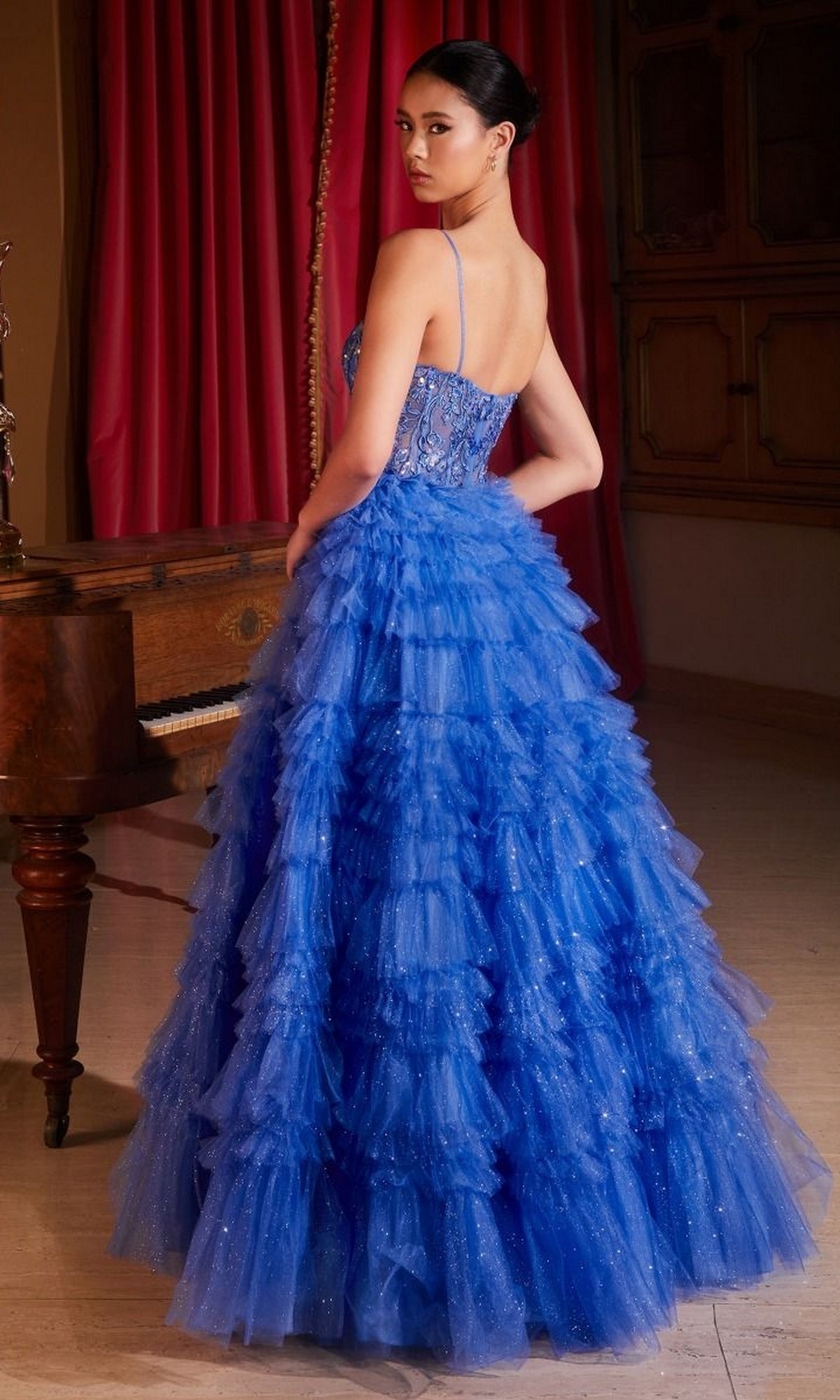 A woman in the Sheer-Corset Long Glitter-Tulle Prom Ball Gown C152 with blue layers and spaghetti straps stands indoors, glancing over her shoulder. A piano and red curtains are seen in the background.