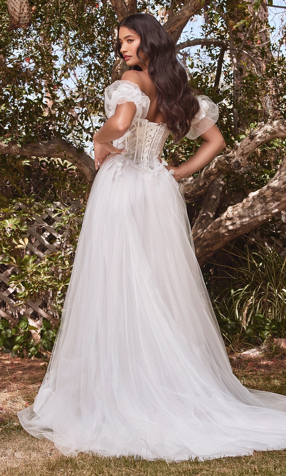 A woman stands outdoors among lush greenery in the Puff-Sleeve Off-White Long Prom Dress CB080W, an off-the-shoulder gown with sheer puff sleeves, a corset back, and a flowing tulle skirt.