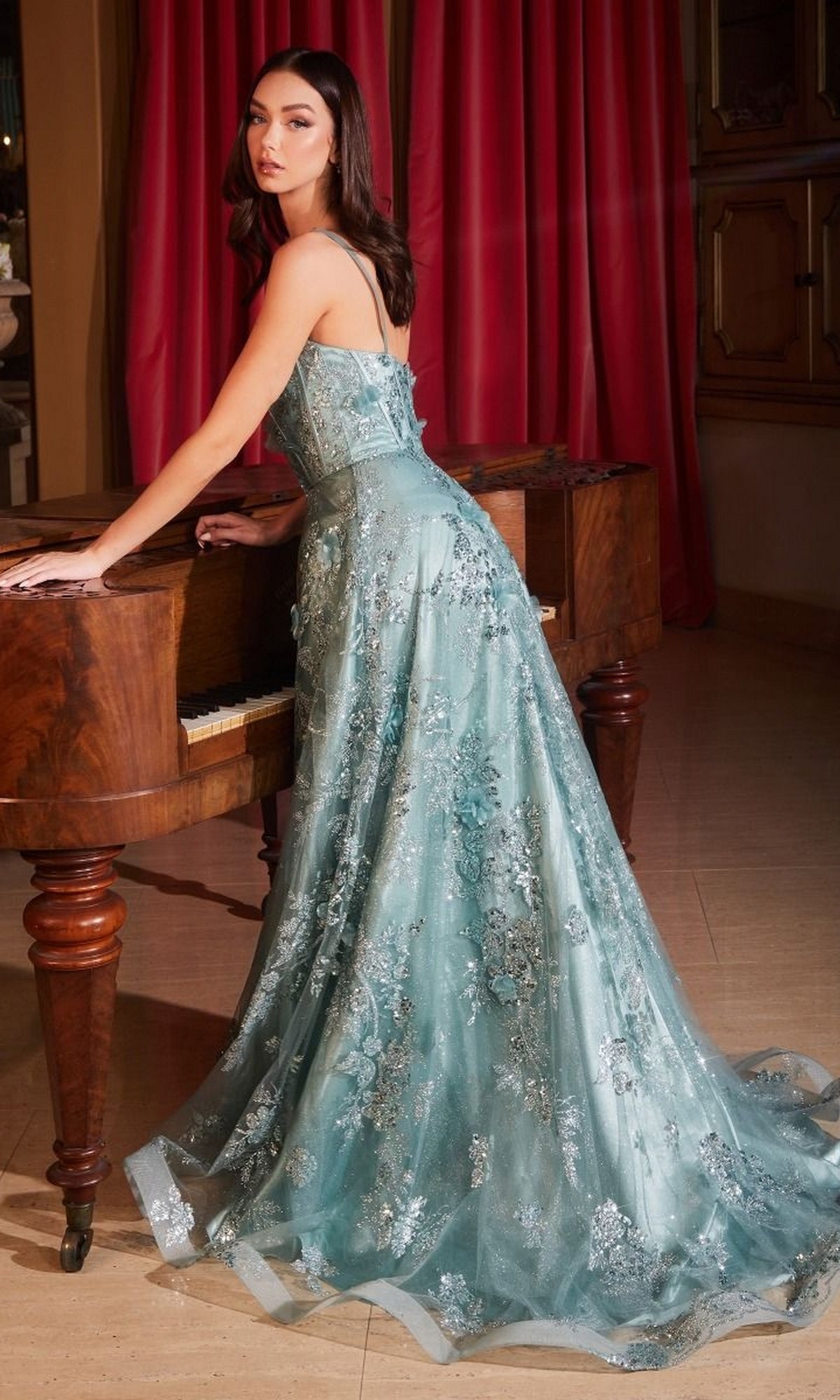 A woman in an elegant room with red curtains stands by a wooden piano, wearing the Long Formal Dress CB144 by Ladivine, a light blue A-line gown with floral appliqué details.