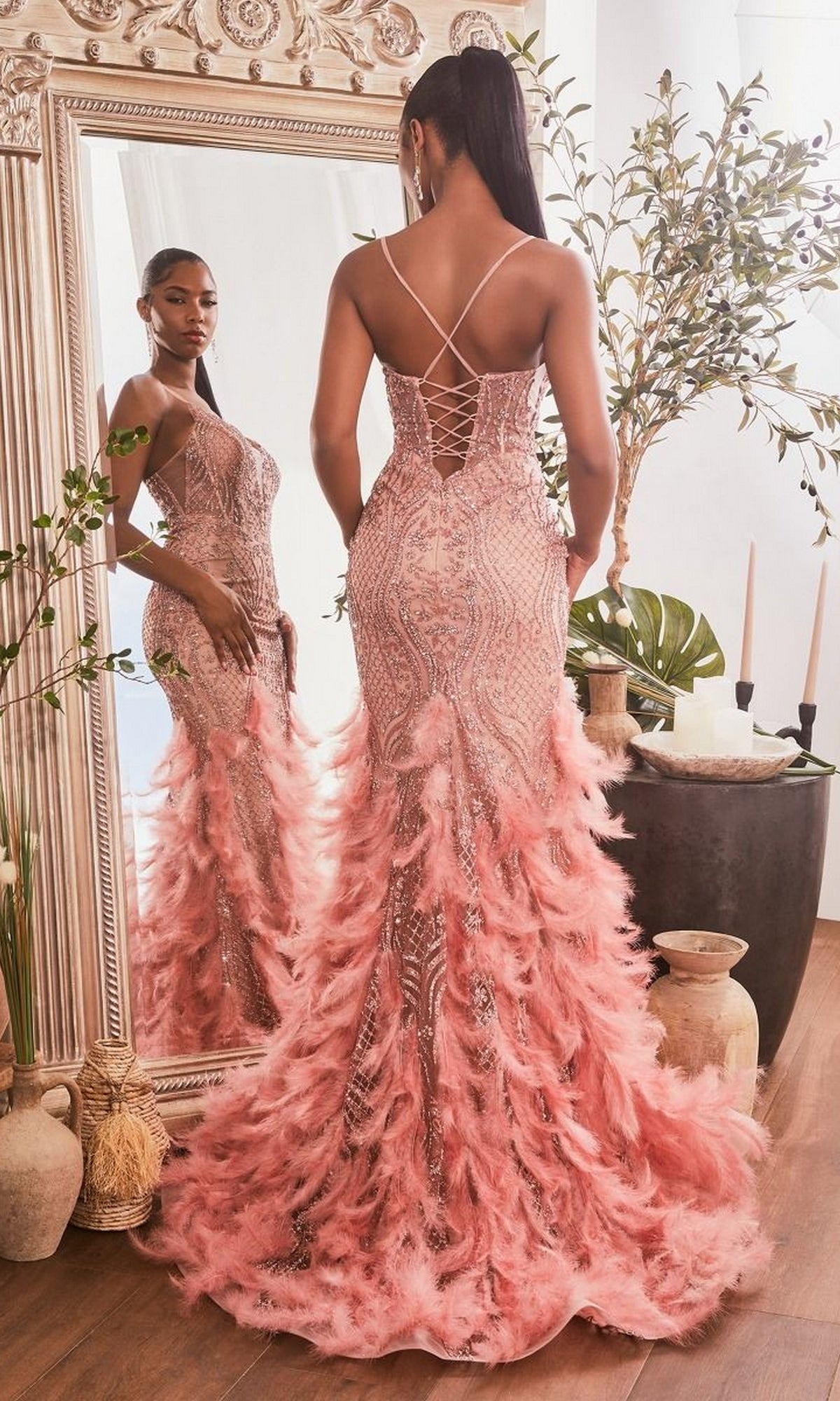A woman in the Feather-Trimmed Glitter-Print Long Prom Dress CC1608 stands before a large ornate mirror, showing off the intricate lace-up back and sweeping train of her stunning pink evening gown.