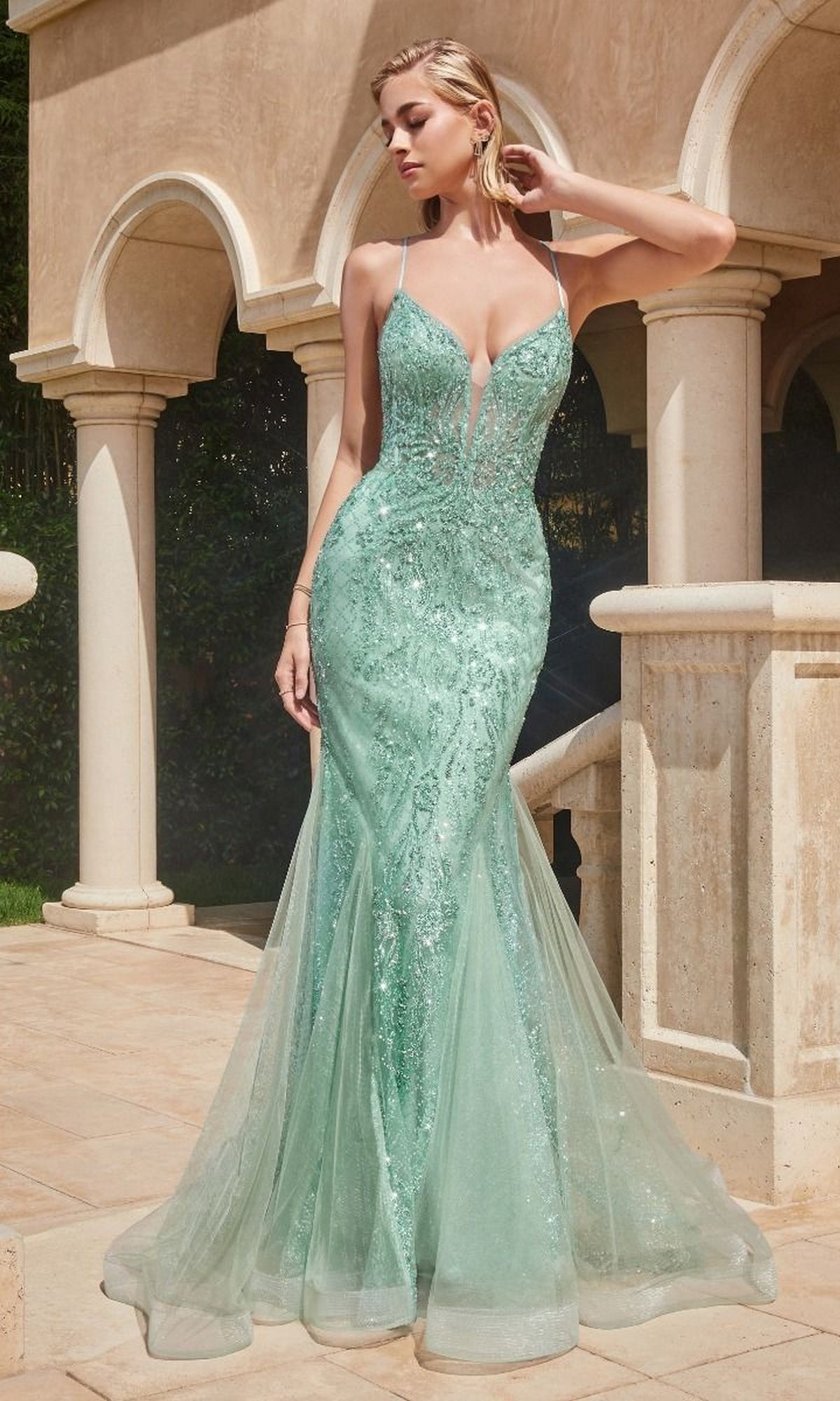 A woman stands outdoors in the Low V-Neck Long Glitter Mermaid Prom Dress CC2253, a fitted mint-green gown with thin straps and a deep V neckline. Stone arches and a staircase are visible in the background.