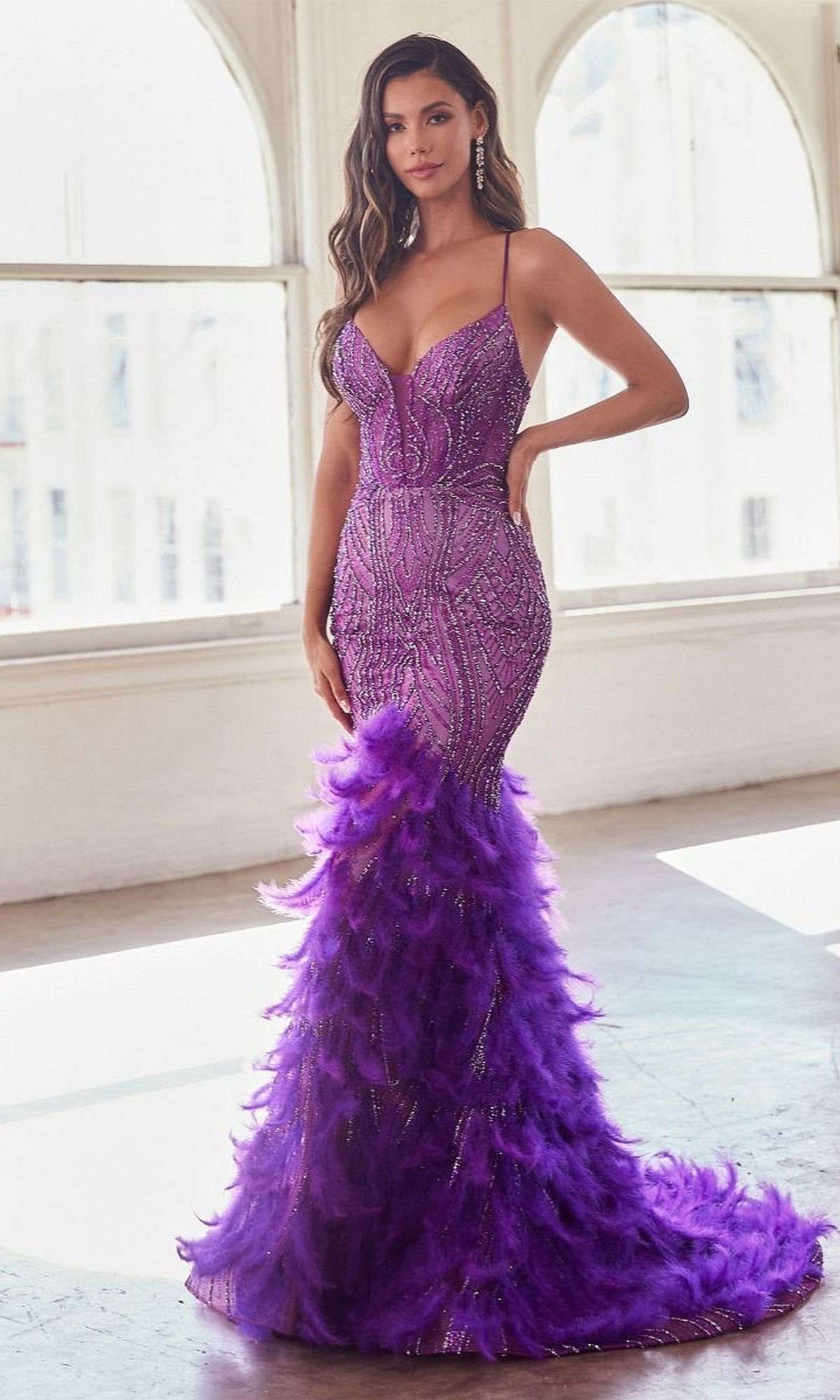 Woman in the Feather-Trim Long Beaded Mermaid Prom Dress CC2308, a fitted purple gown with beading and feathered trim at the hem, stands indoors before large arched windows.
