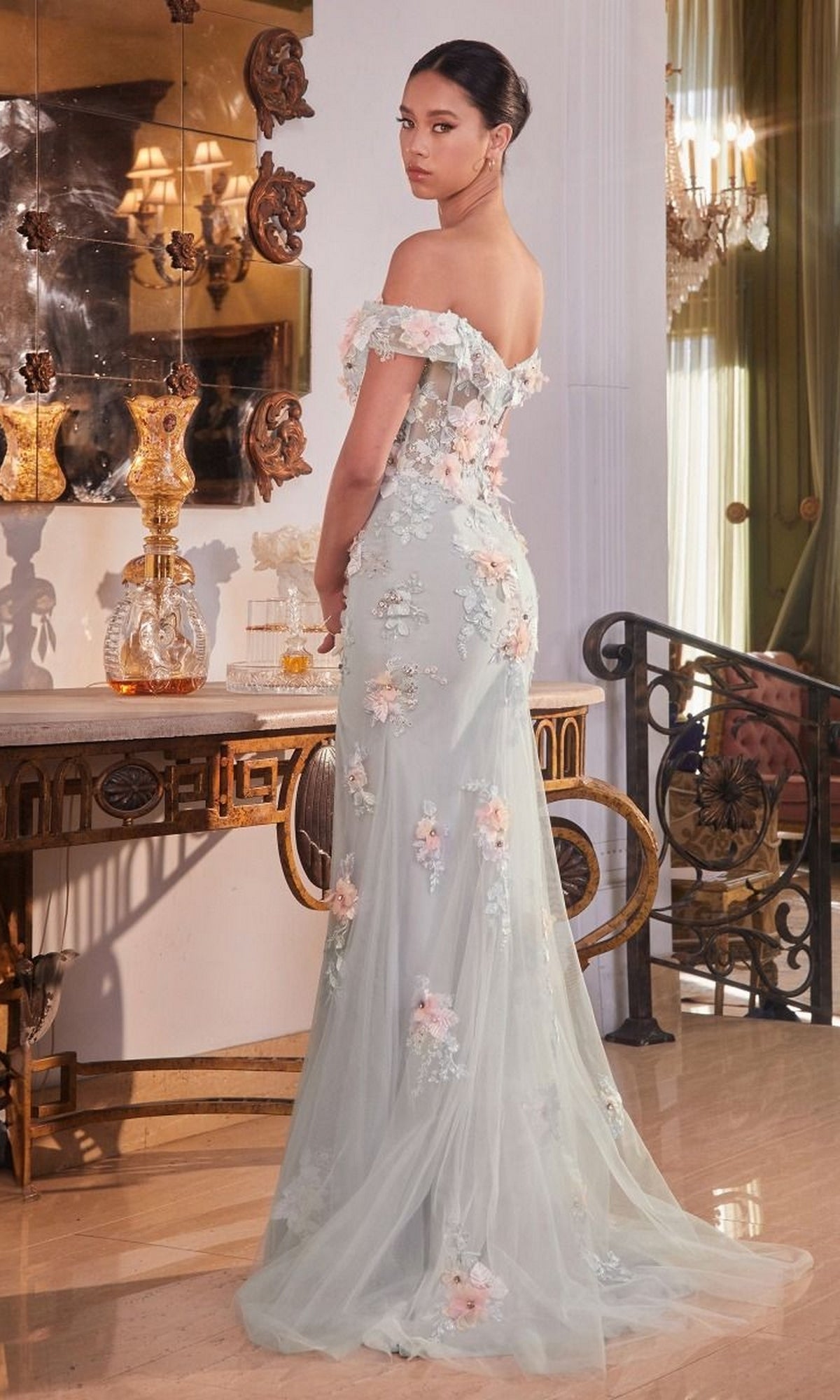A woman poses indoors in the Floral-Embellished Long Mist Gray Prom Dress CD005, featuring a sheer corset bodice, off-the-shoulder sleeves, and delicate floral details. Her pastel gown is turned slightly away from the camera.