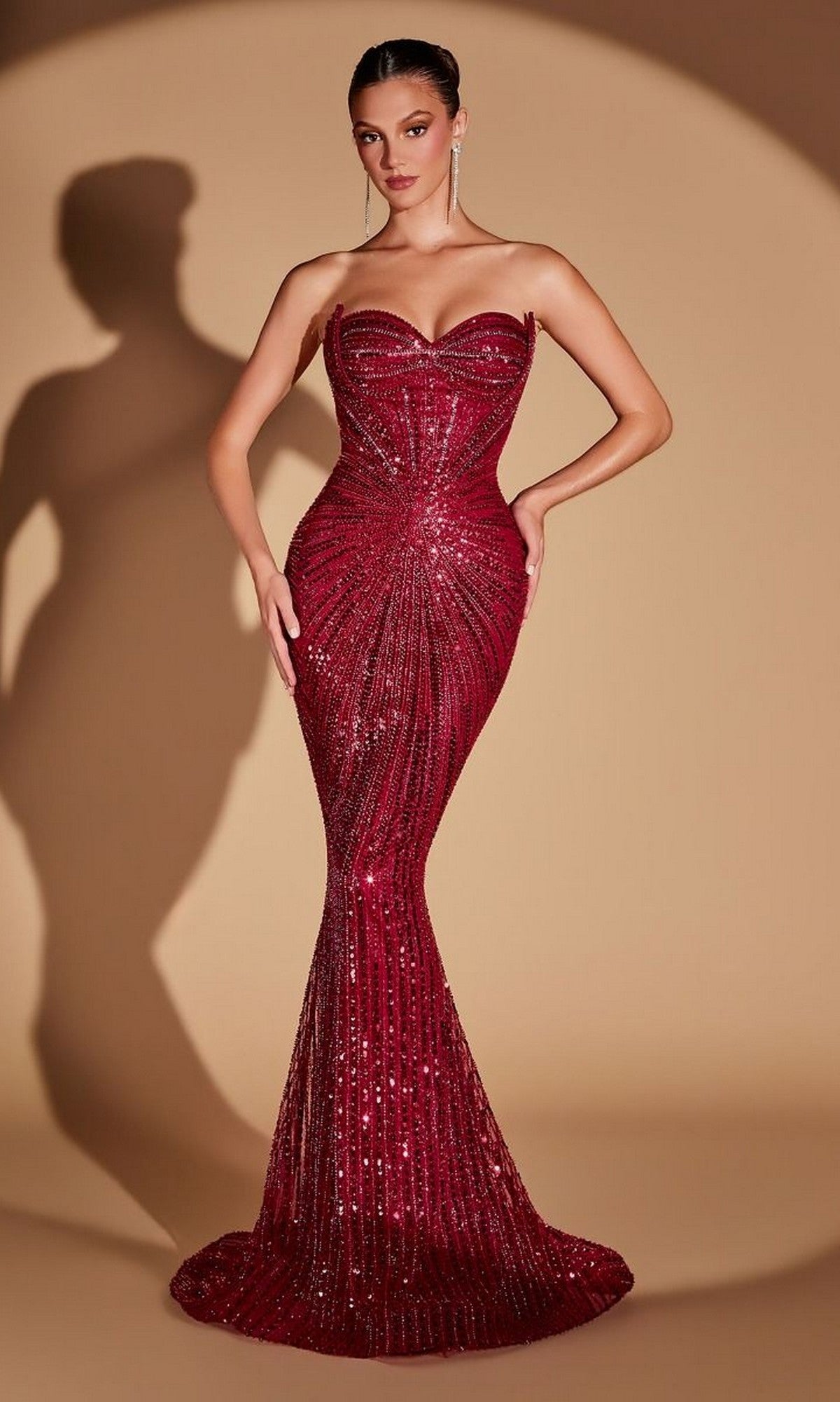 A woman in the strapless, form-fitting Ladivine CD0108 long prom dress in burgundy poses against a beige background with a shadow behind her, the sparkling sequins of her stunning gown catching the light beautifully.