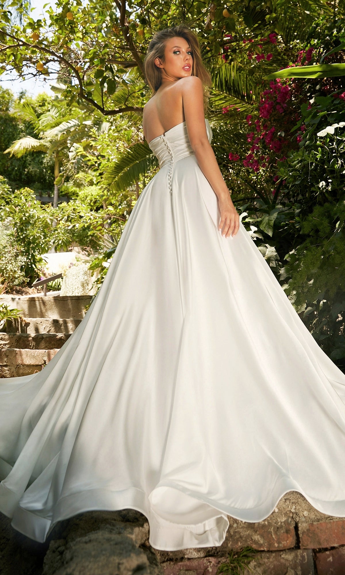 A woman outdoors among greenery and flowers looks over her shoulder, wearing the Strapless Long White Satin Formal Dress CD0166W with a flowing train.