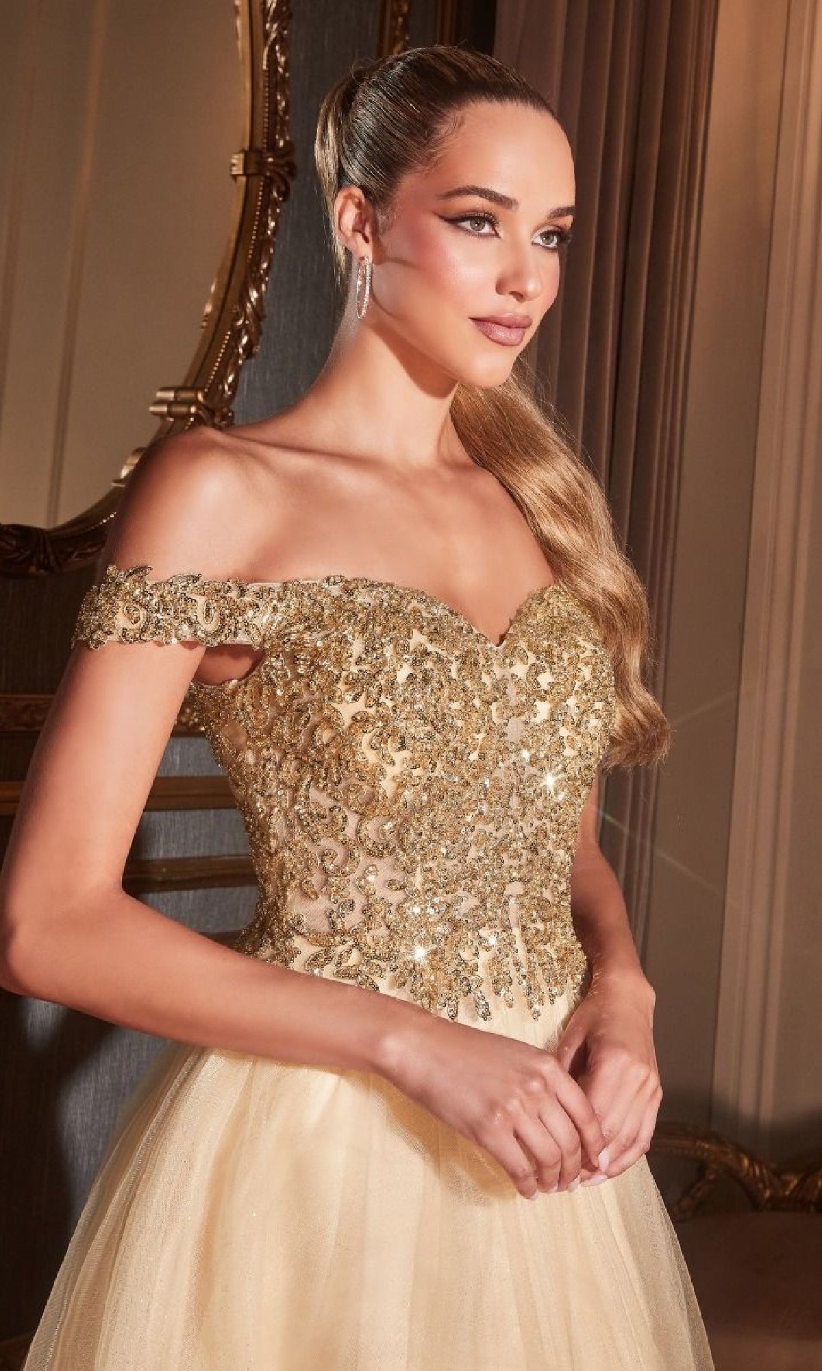A woman in the Beaded-Bodice Long Prom Dress: Ladivine CD0177 stands indoors, wearing an off-the-shoulder gown with gold embellishments, hands folded and gazing to the side.