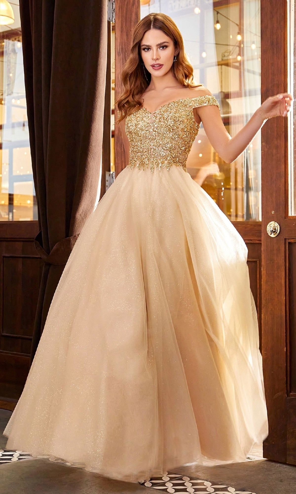 A woman in the Ladivine CD0177 Beaded-Bodice Long Prom Dress with a glittery fitted bodice and full tulle skirt stands indoors by wooden doors and brown curtains.