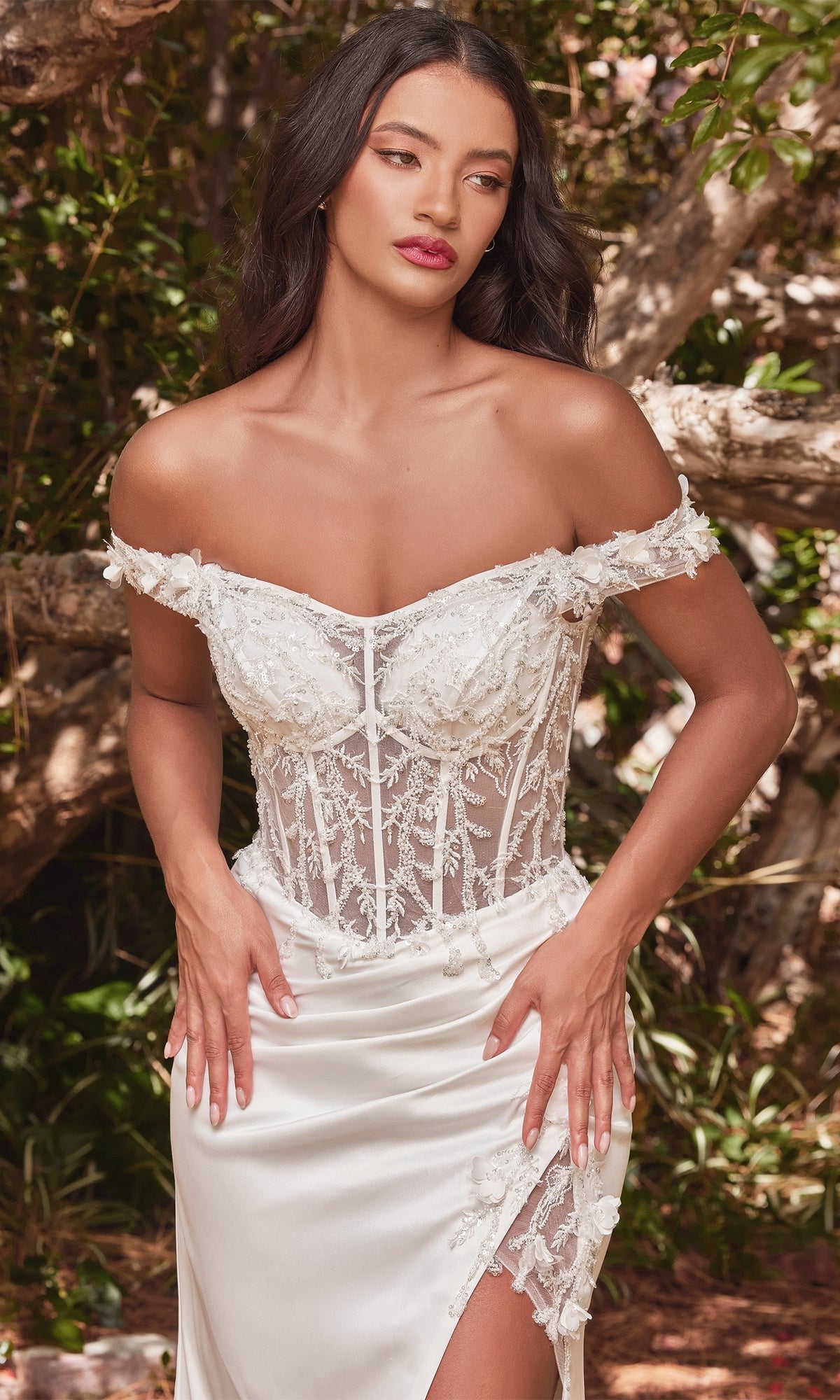 A woman stands outdoors near trees in the Off-Shoulder Long White Corset Prom Dress CD0186W, featuring white lace, a fitted bodice, and a thigh-high slit for an elegant, evening-inspired bridal look.