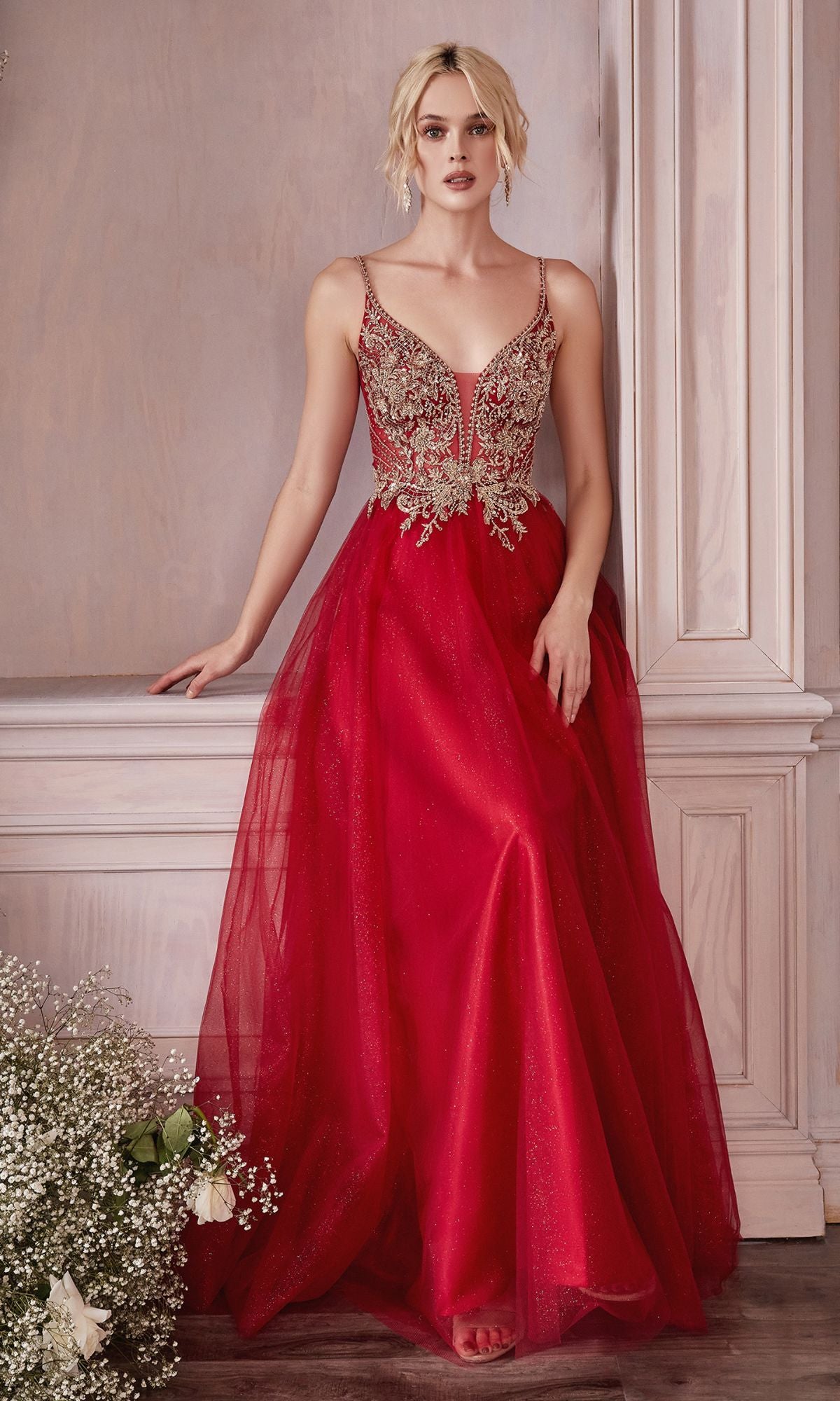 A woman in the Embellished-Bodice Long Tulle Prom Dress CD0195, featuring a red sheer skirt and gold embroidered bodice, stands against a cream wall next to white flowers.