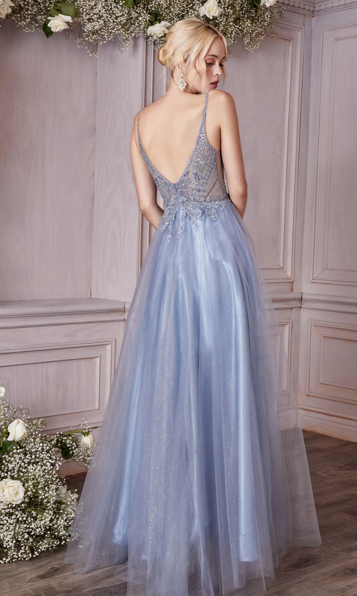 A woman in the affordable Embellished-Bodice Long Tulle Prom Dress CD0195, a light blue backless a-line gown with lace detail, stands before a wooden wall adorned with white flowers and greenery.