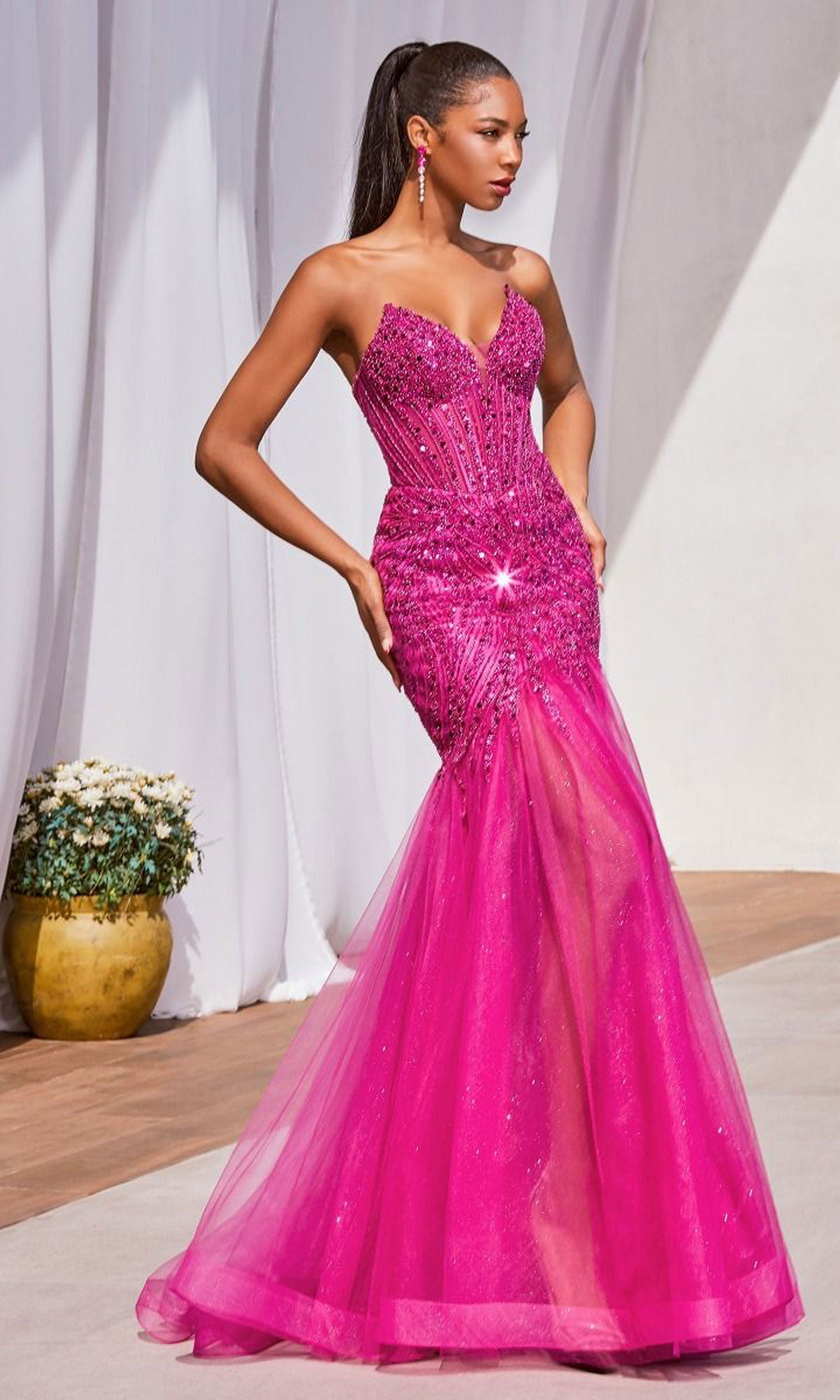 A woman stands outdoors near white drapery and a potted plant, wearing the Strapless Beaded Long Mermaid Prom Dress CD0214 in hot pink with a beaded bodice and long tulle skirt.