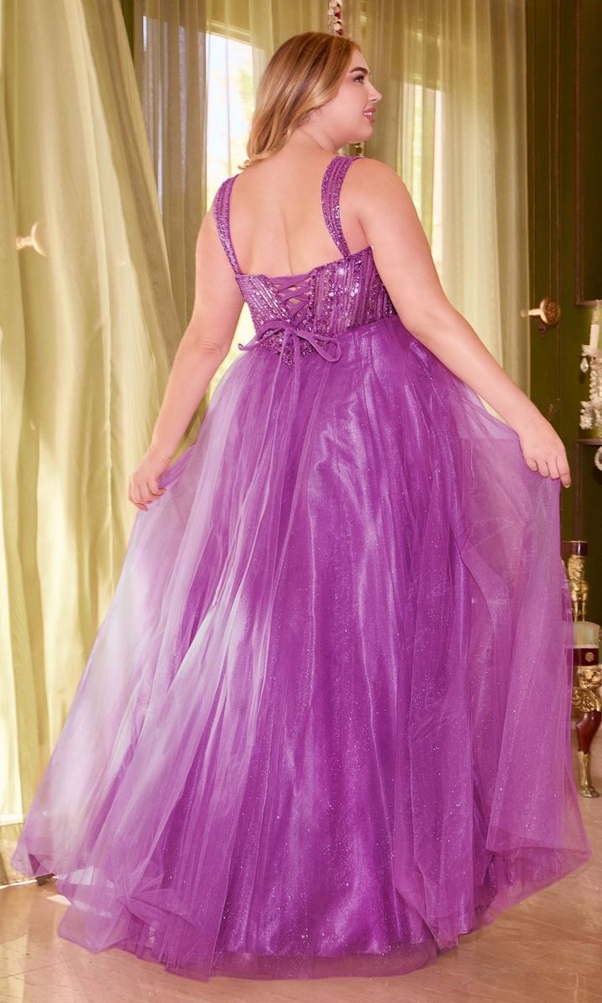 A woman stands indoors wearing the Plus-Size Sequin-Bodice Long Prom Dress CD0217C—a sleeveless, floor-length purple gown with a fitted sequin bodice and a flowing tulle skirt, shown from the back.