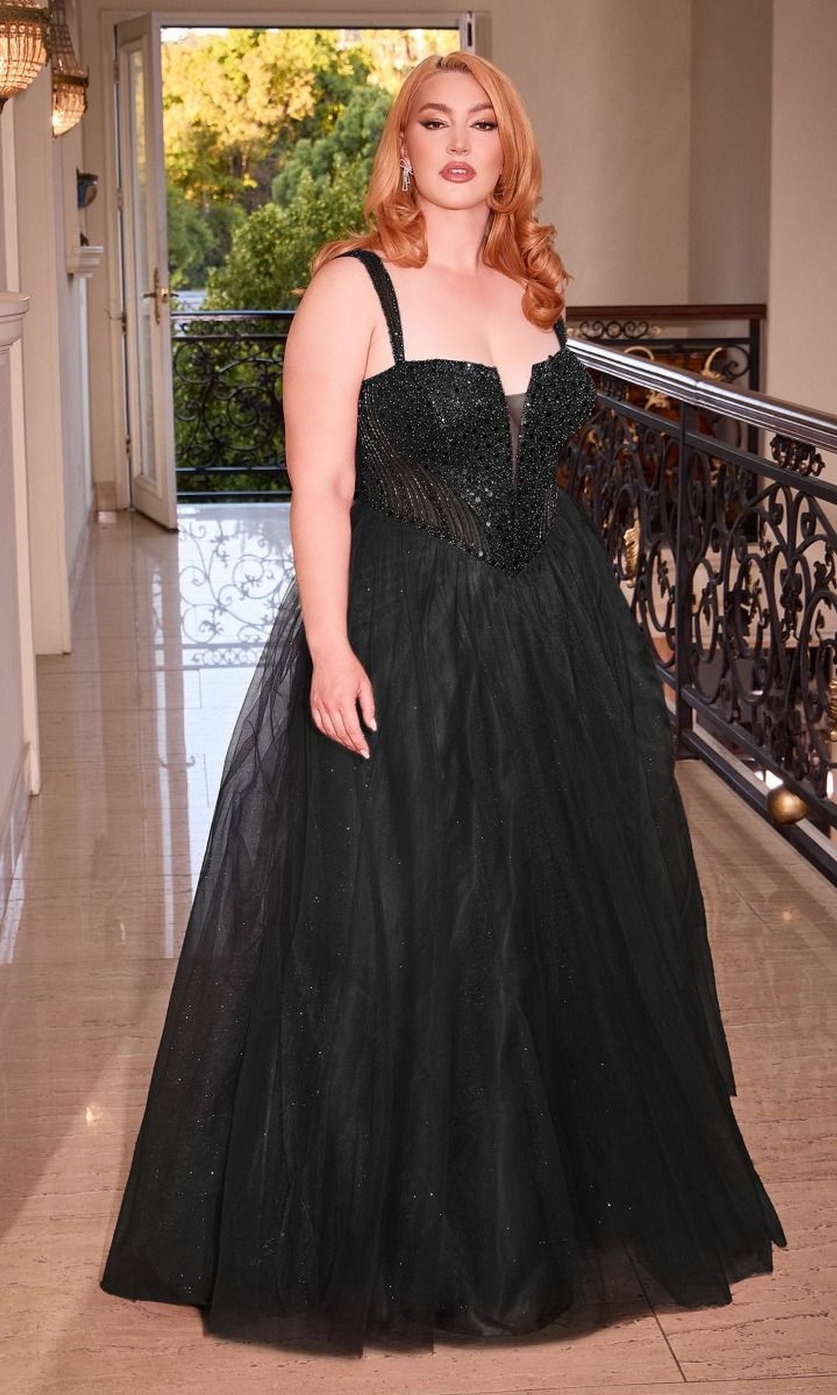 A woman with long red hair stands indoors, wearing the Plus-Size Sequin-Bodice Long Prom Dress CD0217C—a floor-length black gown with a fitted sequin bodice and tulle skirt. A balcony and greenery are visible through an open door behind her.