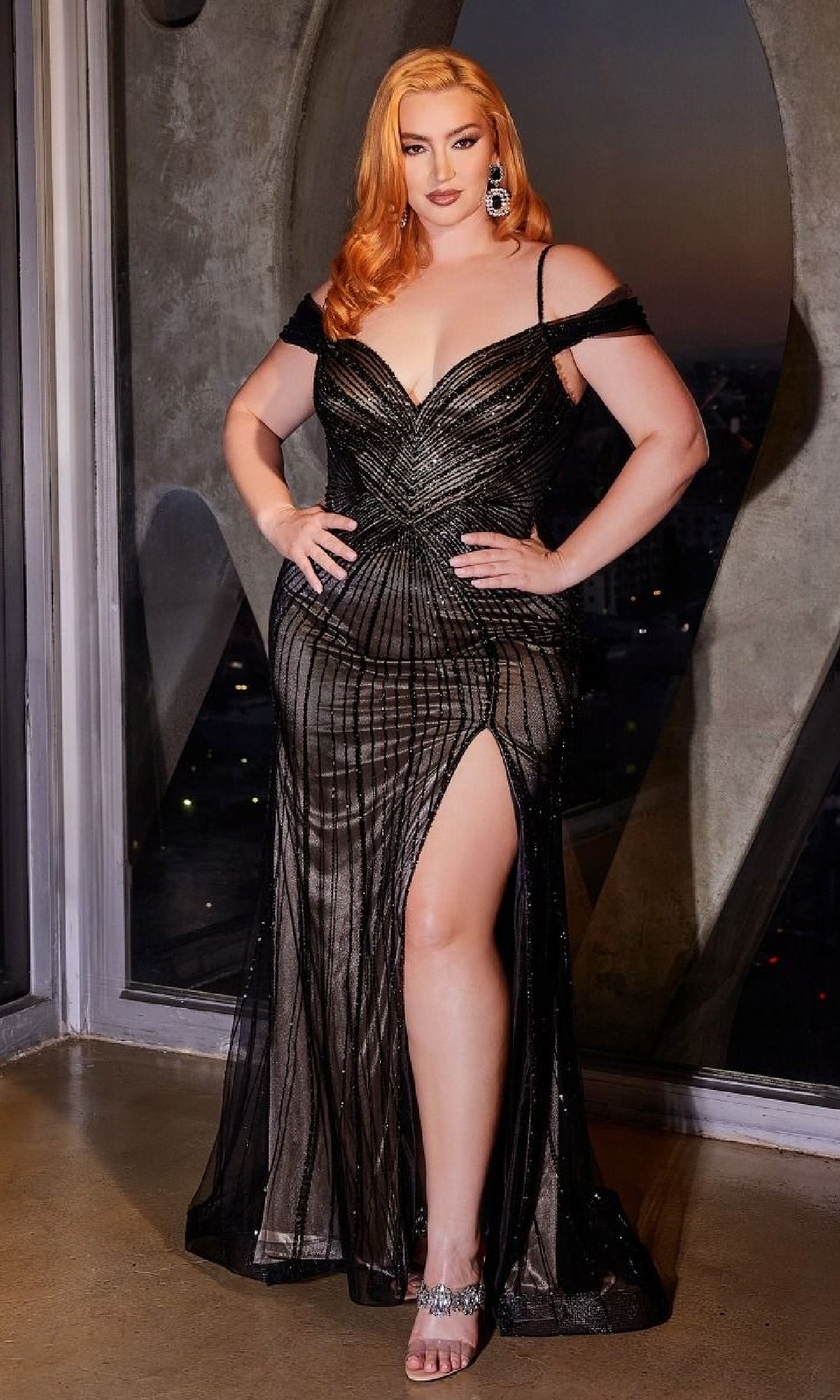 A red-haired woman poses indoors at sunset, wearing the Plus-Size Beaded Long Prom Dress Ladivine CD0219C, a black off-the-shoulder gown with a thigh-high slit and silver heels.
