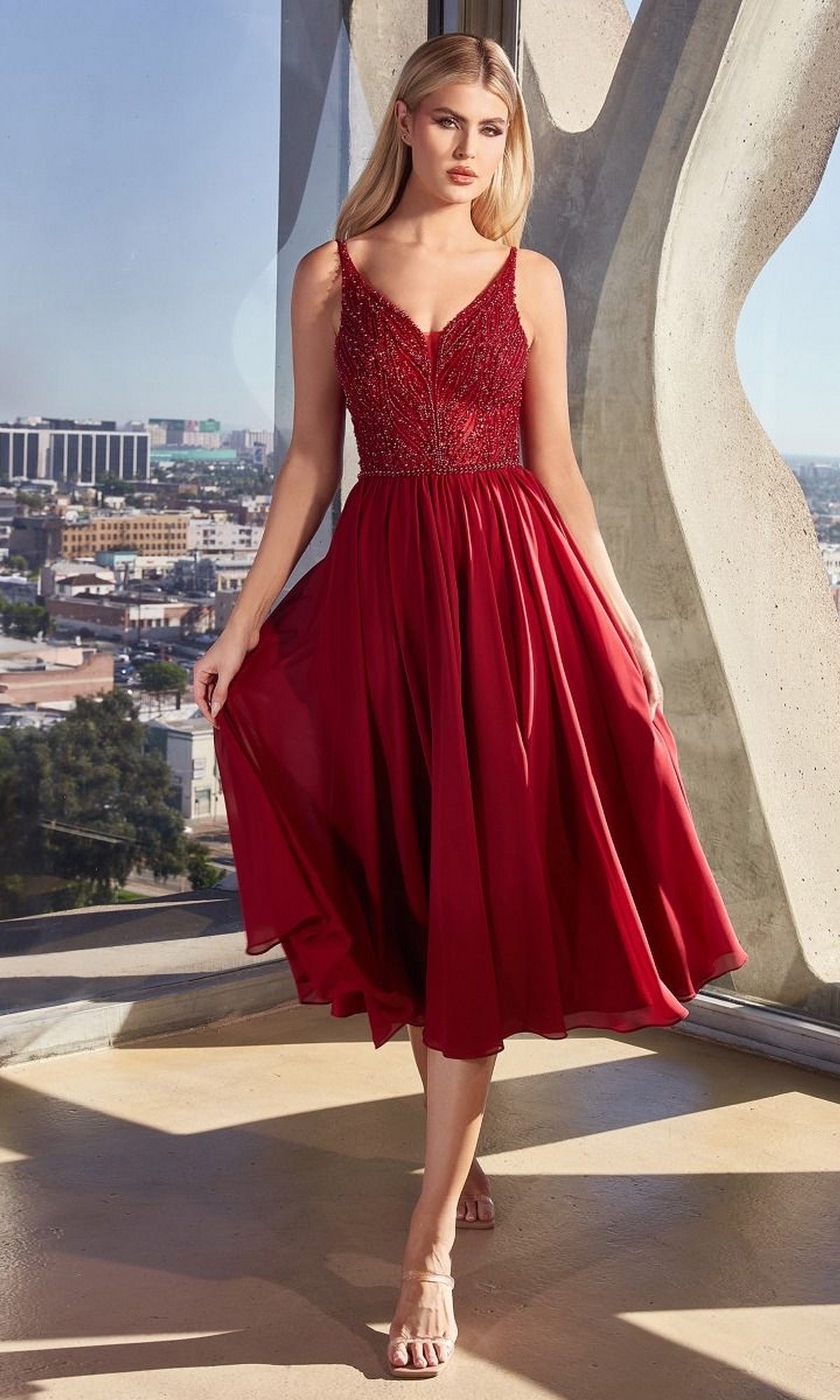 A woman in the Sheer-Bodice Tea-Length A-Line Party Dress CD0225, deep red with a beaded bodice and flowing skirt, stands indoors by a large window overlooking the city.