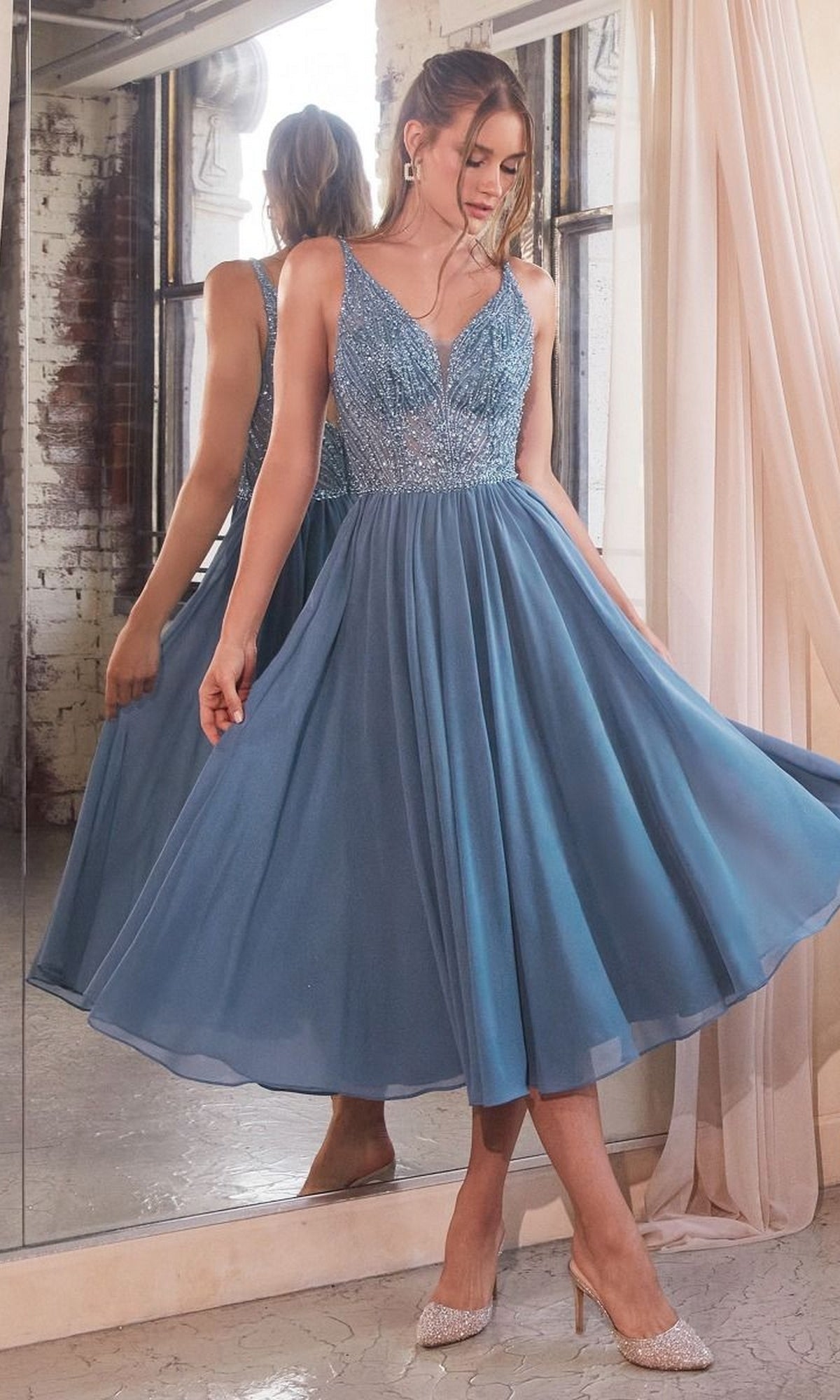 A woman in the Sheer-Bodice Tea-Length A-Line Party Dress CD0225 stands indoors by a window, showcasing the skirt of her blue semi-formal dress with a beaded bodice.