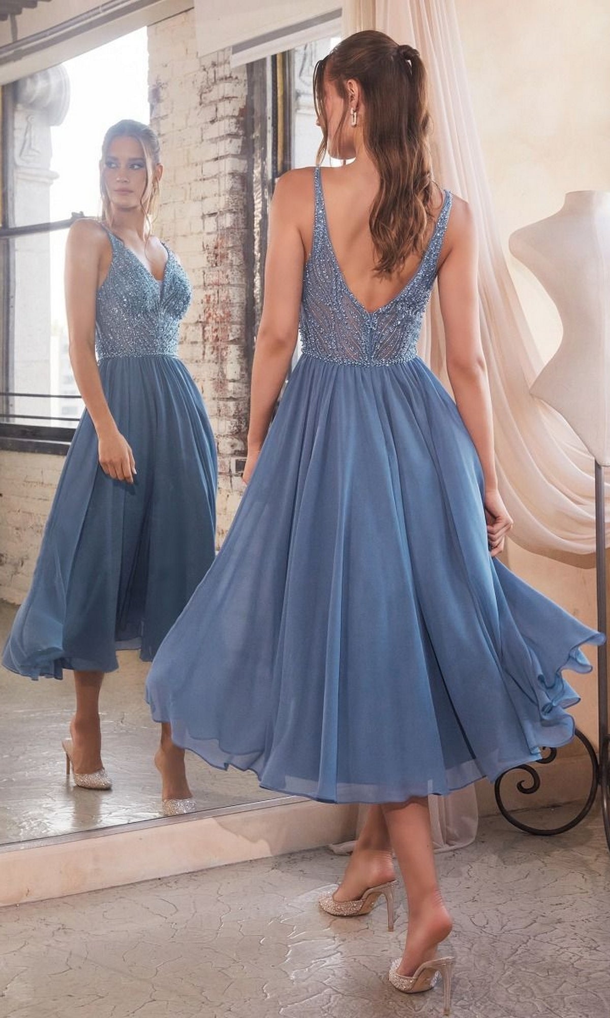 A woman in the Sheer-Bodice Tea-Length A-Line Party Dress CD0225, featuring a blue beaded bodice and flared skirt, stands before a mirror holding her shoes.
