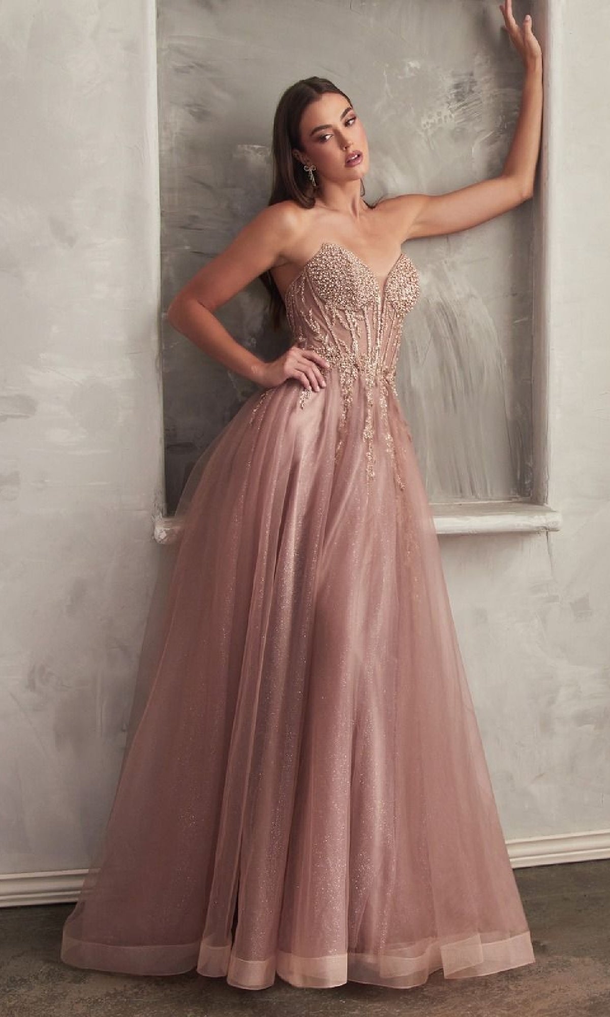 A woman poses elegantly against a textured gray wall in the Strapless Sheer-Corset Long Prom Ball Gown CD0230, featuring a blush pink hue, fitted embellished bodice, and flowing glitter tulle skirt.