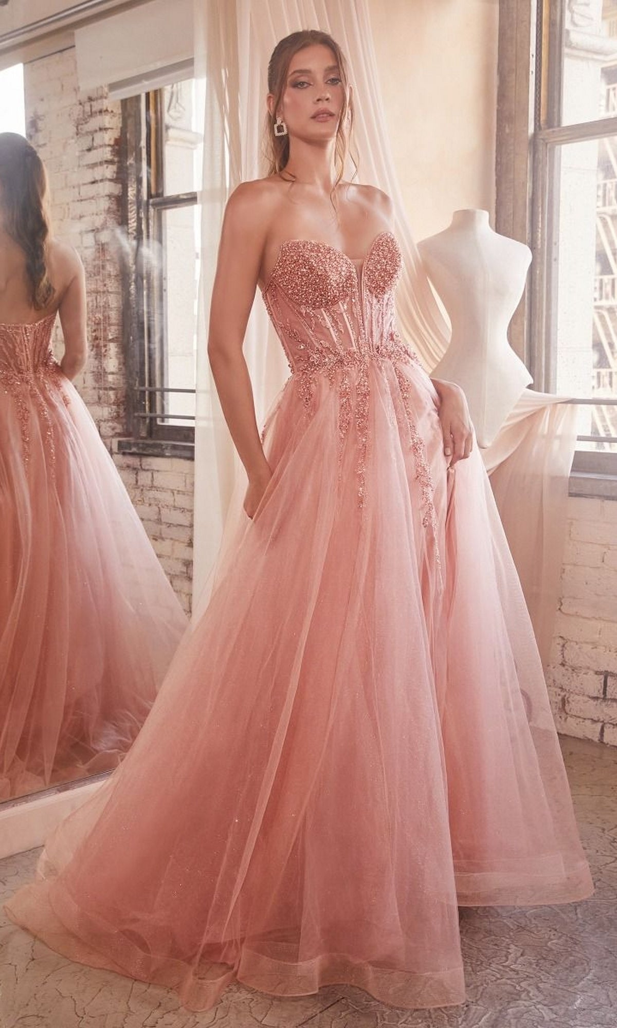 A woman in the Strapless Sheer-Corset Long Prom Ball Gown CD0230—a floor-length blush pink glitter tulle dress with a fitted embellished bodice—stands indoors by a window and dress form.