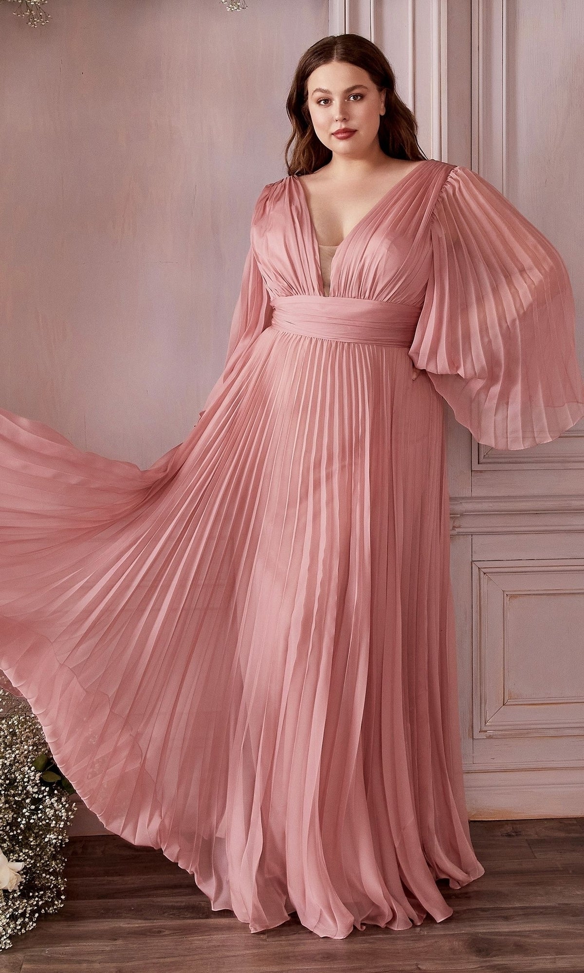 A woman in the Long Sleeve Plus-Size Long Formal Dress CD242C, featuring a flowing rose pink pleated design and sheer balloon sleeves, stands in a softly lit room with wooden floors and light wall paneling.