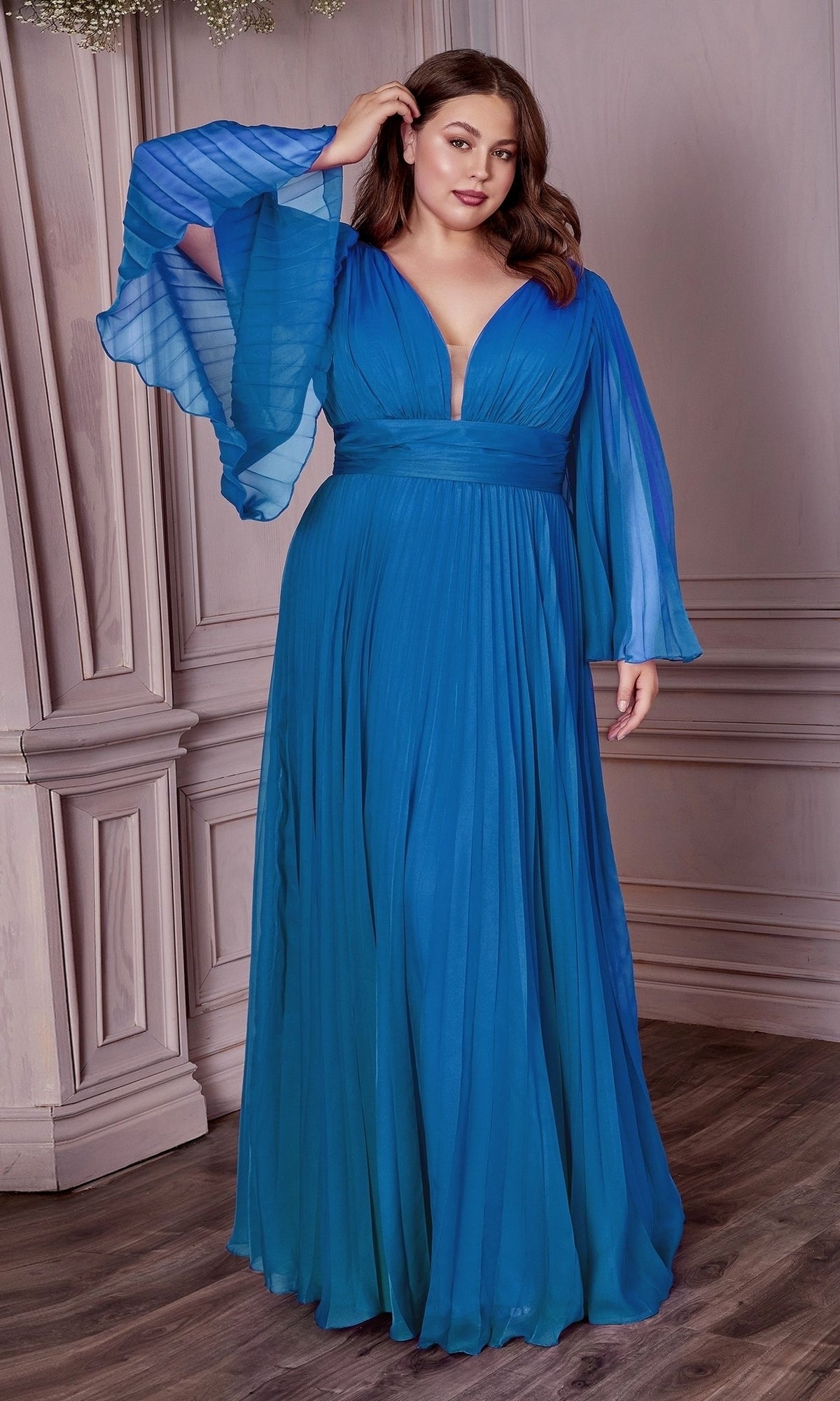 A woman stands indoors against a decorative paneled wall, wearing the Long Sleeve Plus-Size Long Formal Dress CD242C—a flowing evening gown with sheer, wide sleeves.