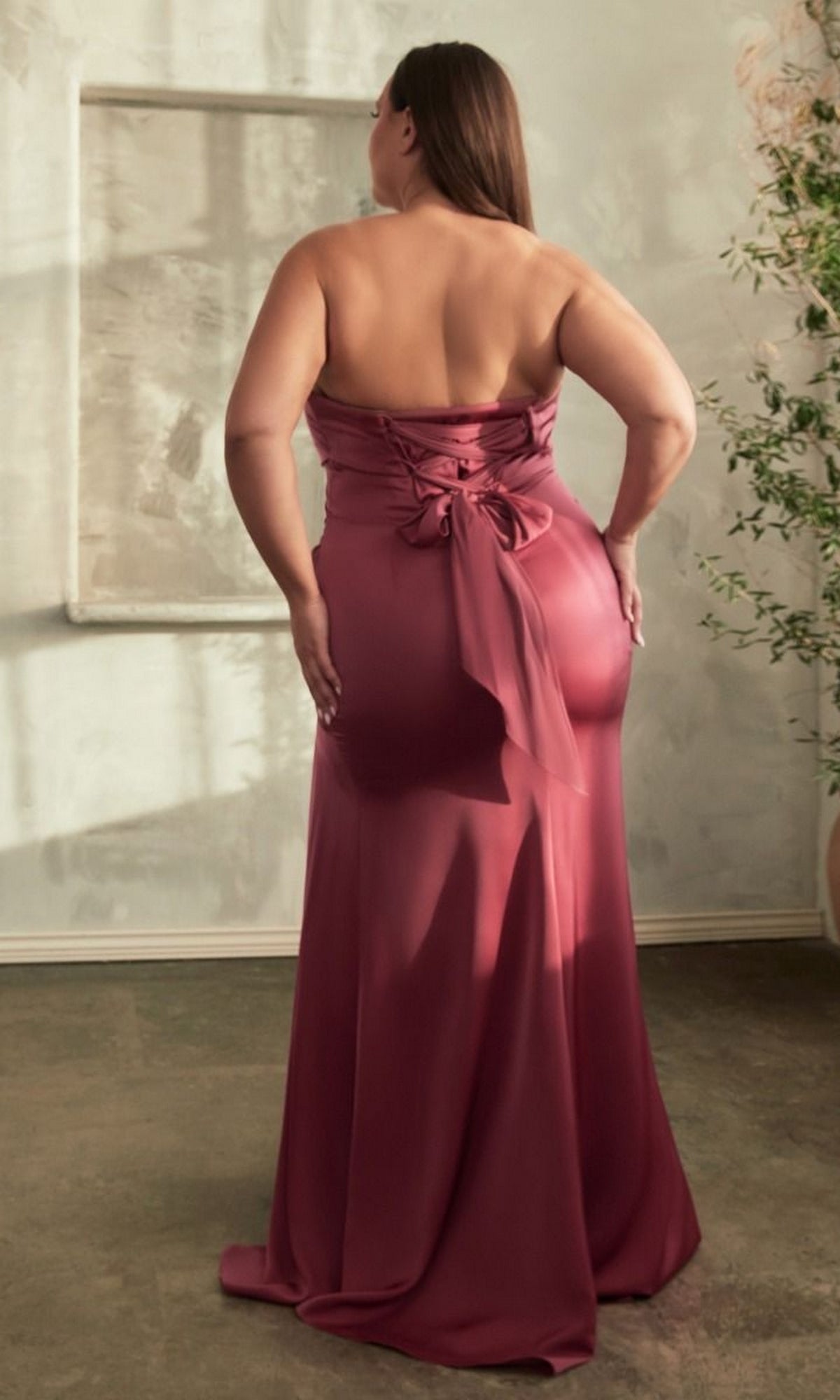 A woman in the Lace-Up Plus-Size Strapless Long Prom Dress CD326C stands with her back to the camera in a softly lit room, showcasing the gown's mauve hue, corset-style bodice, lace-up back, and delicate bow detail.