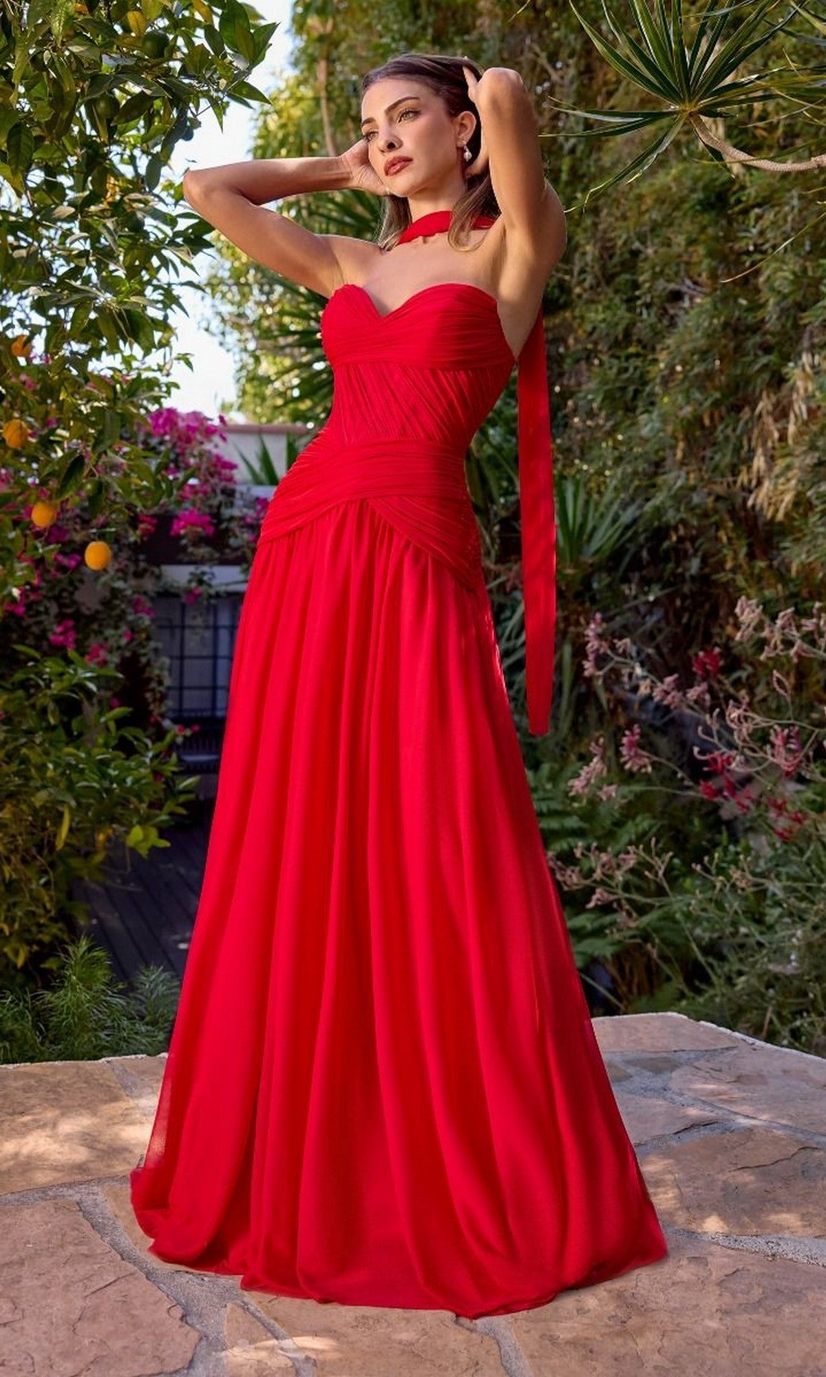 A woman stands outdoors in the Ladivine CD395, a long, strapless red prom dress with a corset bodice and matching neck scarf, surrounded by lush greenery and colorful flowers.