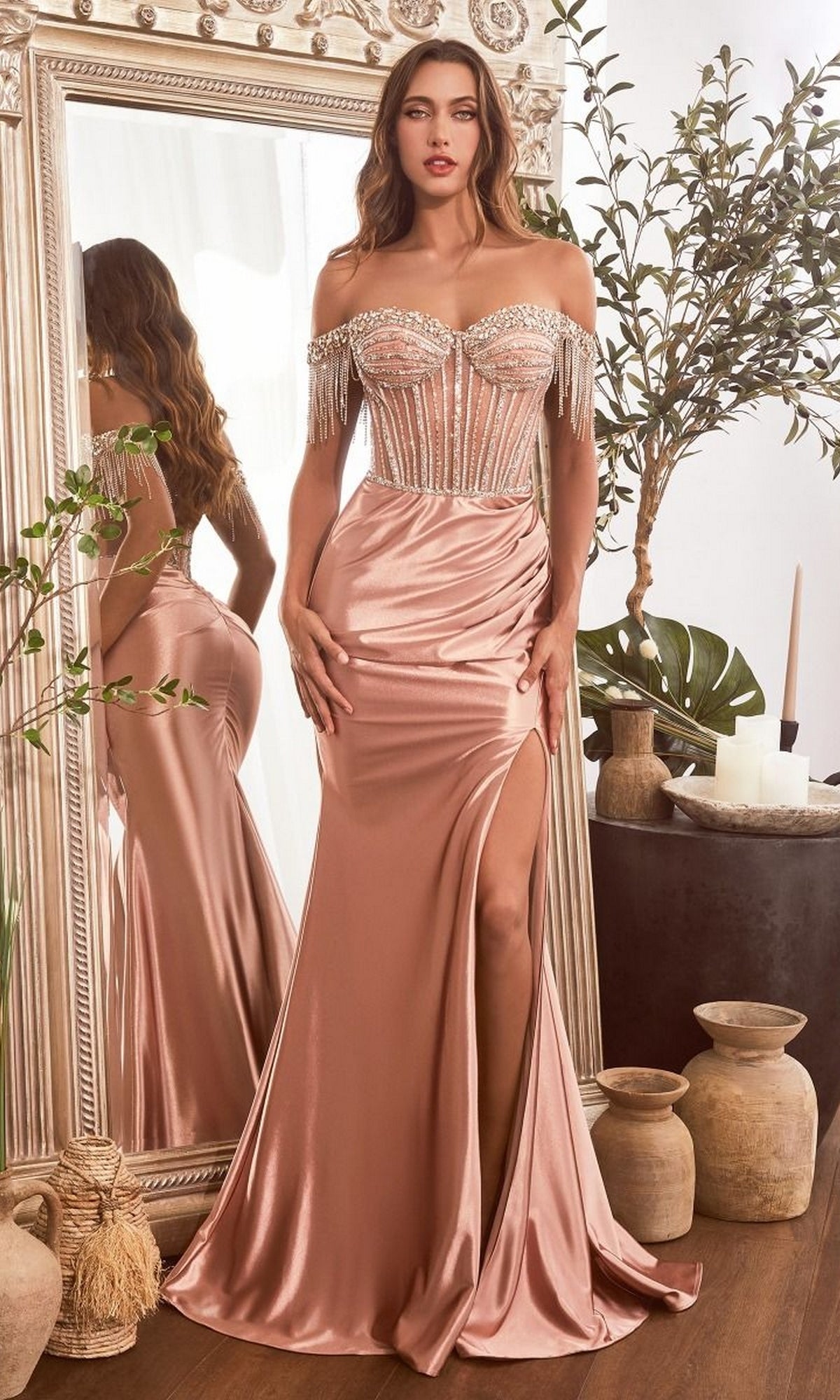 A woman in the Beaded-Fringe Long Prom Gown: Ladivine CD821, a pink satin off-the-shoulder dress with beaded corset and thigh-high slit, stands before an ornate mirror surrounded by decorative vases and lush plants.